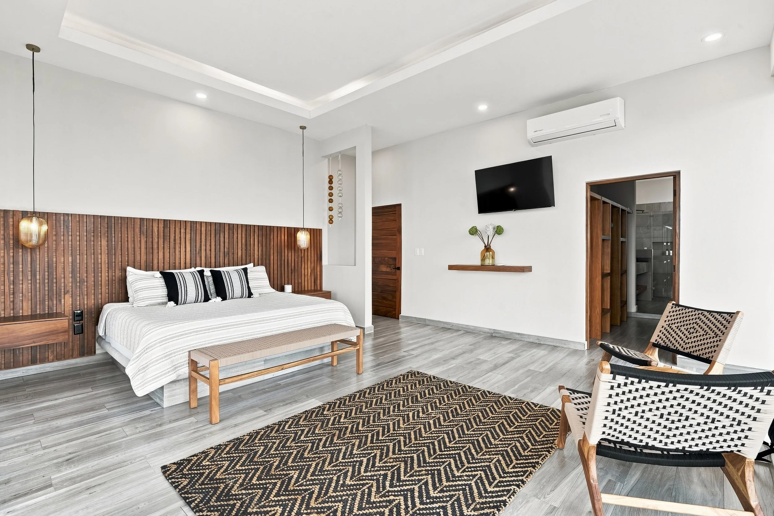 Habitación moderna con cama de madera, 2 sillas de mimbre con estructura de madera y respaldo de cuerda, alfombra con patron de zigzag, paredes blancas, lámparas de techo, televisor y aire acondicionado.