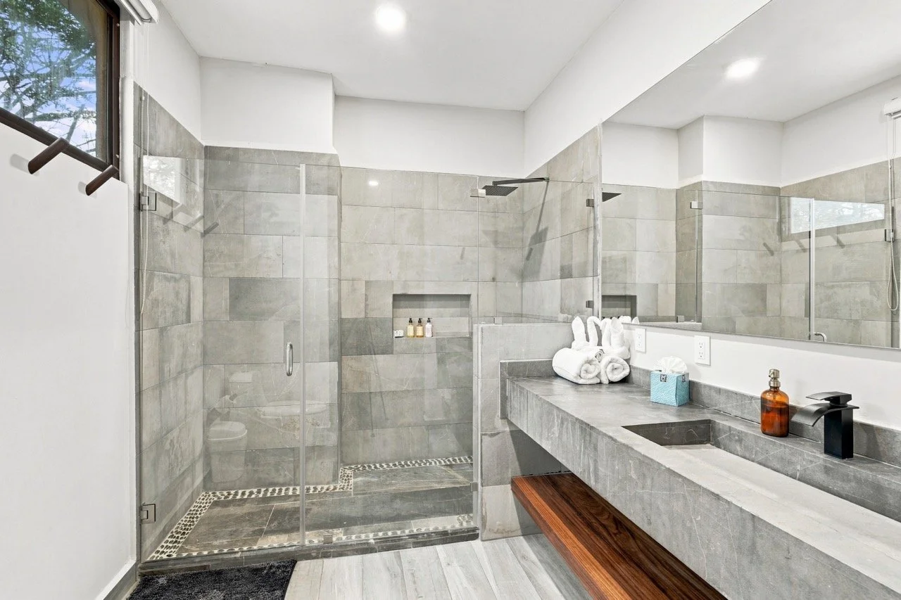 Baño moderno con ducha de cristal, lavabo de piedra con dispensador de jabón y toallas dobladas, y adornos de spa, en tonos grises y blancos.