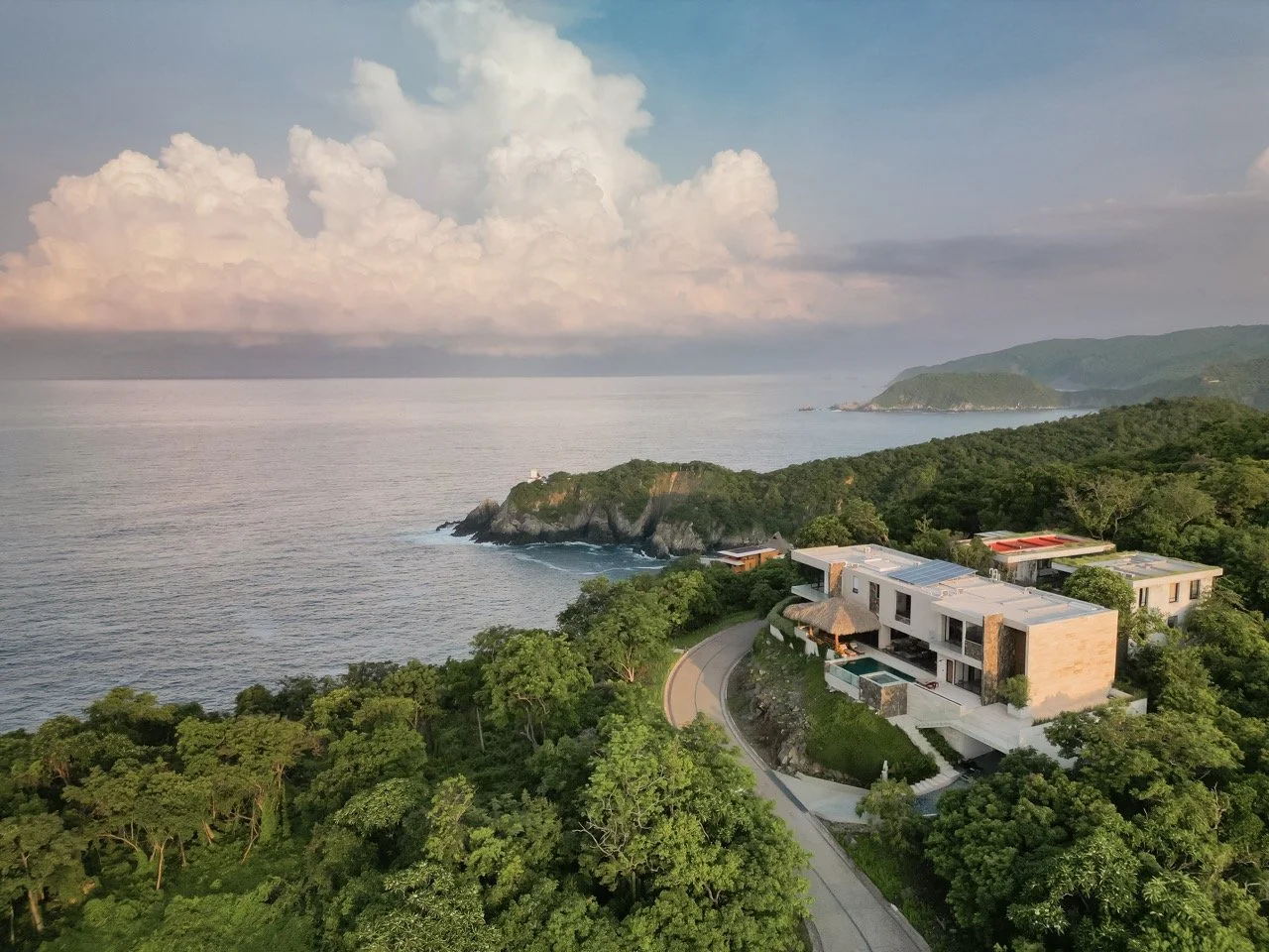 Vista aérea de una playa con agua tranquila, acantilados verdes y un conjunto de casas modernas rodeadas de árboles, con colinas al fondo y cielo nublado.