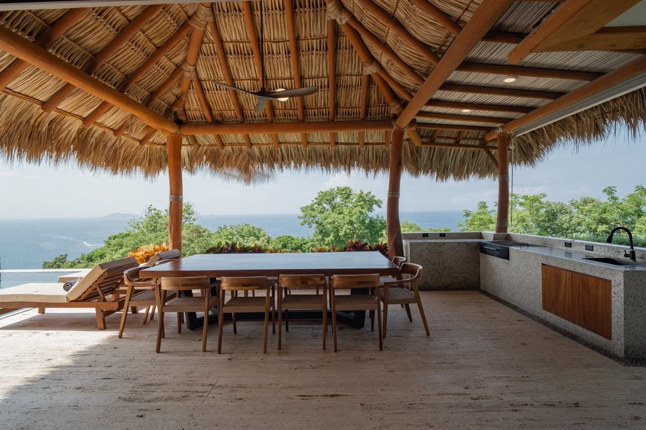 Áreas de comedor y cocina al aire libre con vista al mar y a la naturaleza en un palapa con techo de paja.