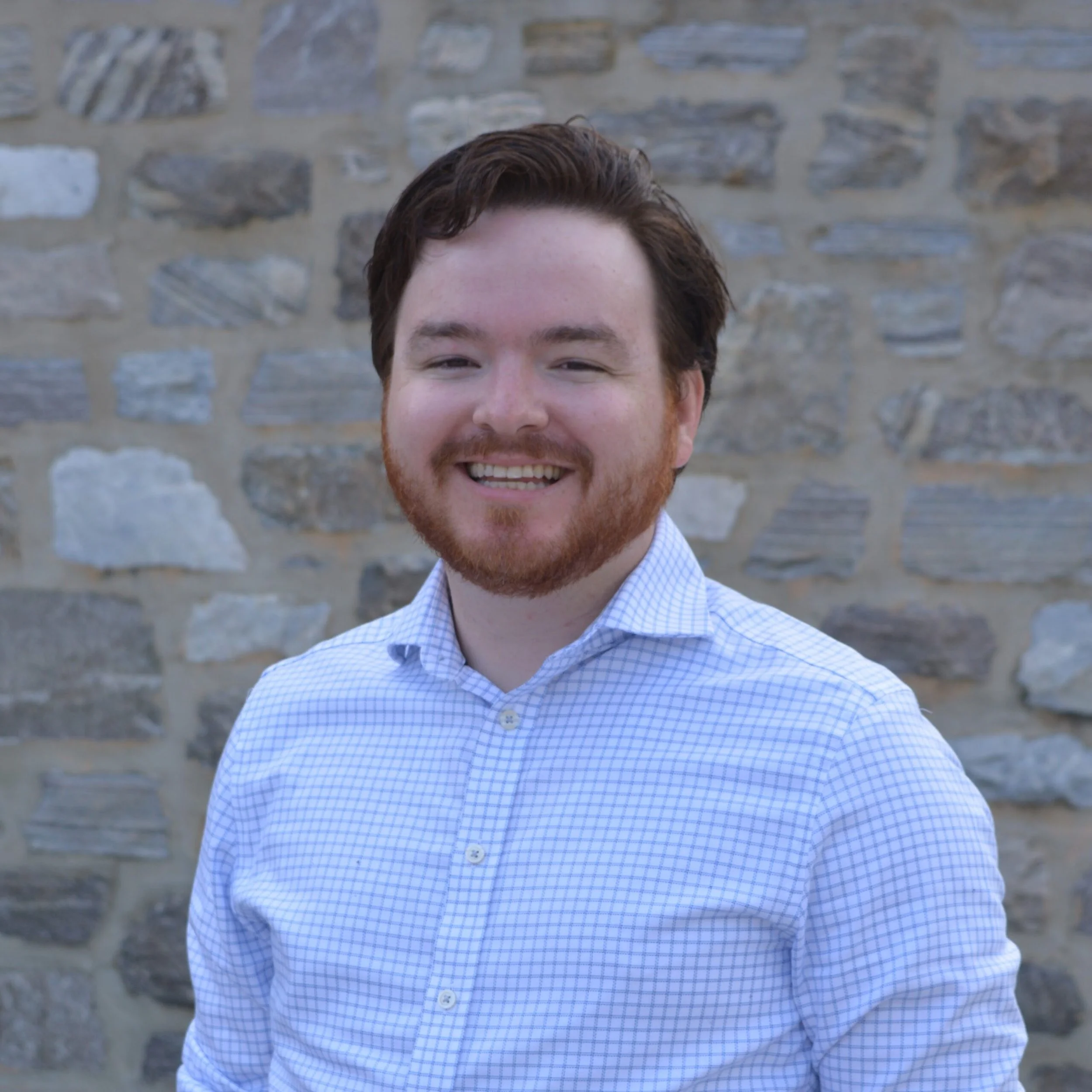 Close-up of a smiling young man with brown hair and a reddish beard, wearing a light blue checkered shirt, standing against a stone wall background.