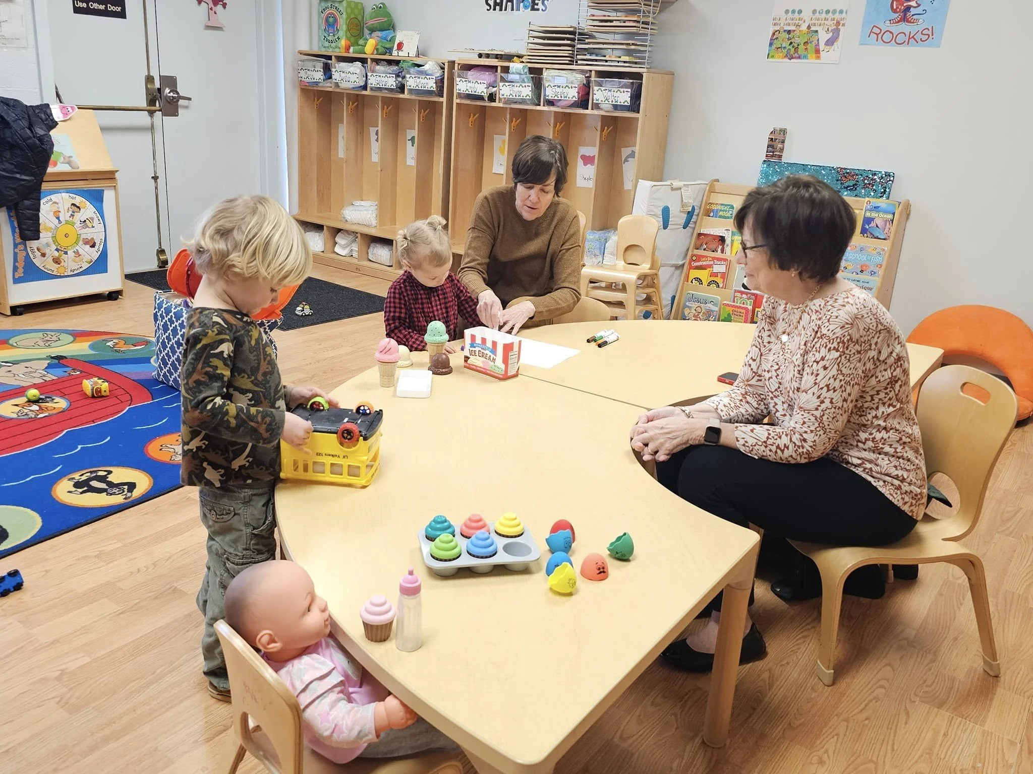 A classroom setting with children and adults sitting around a table engaging in a play activity with toys. The room has colorful decorations, shelves with bins, and children's artwork on the walls.