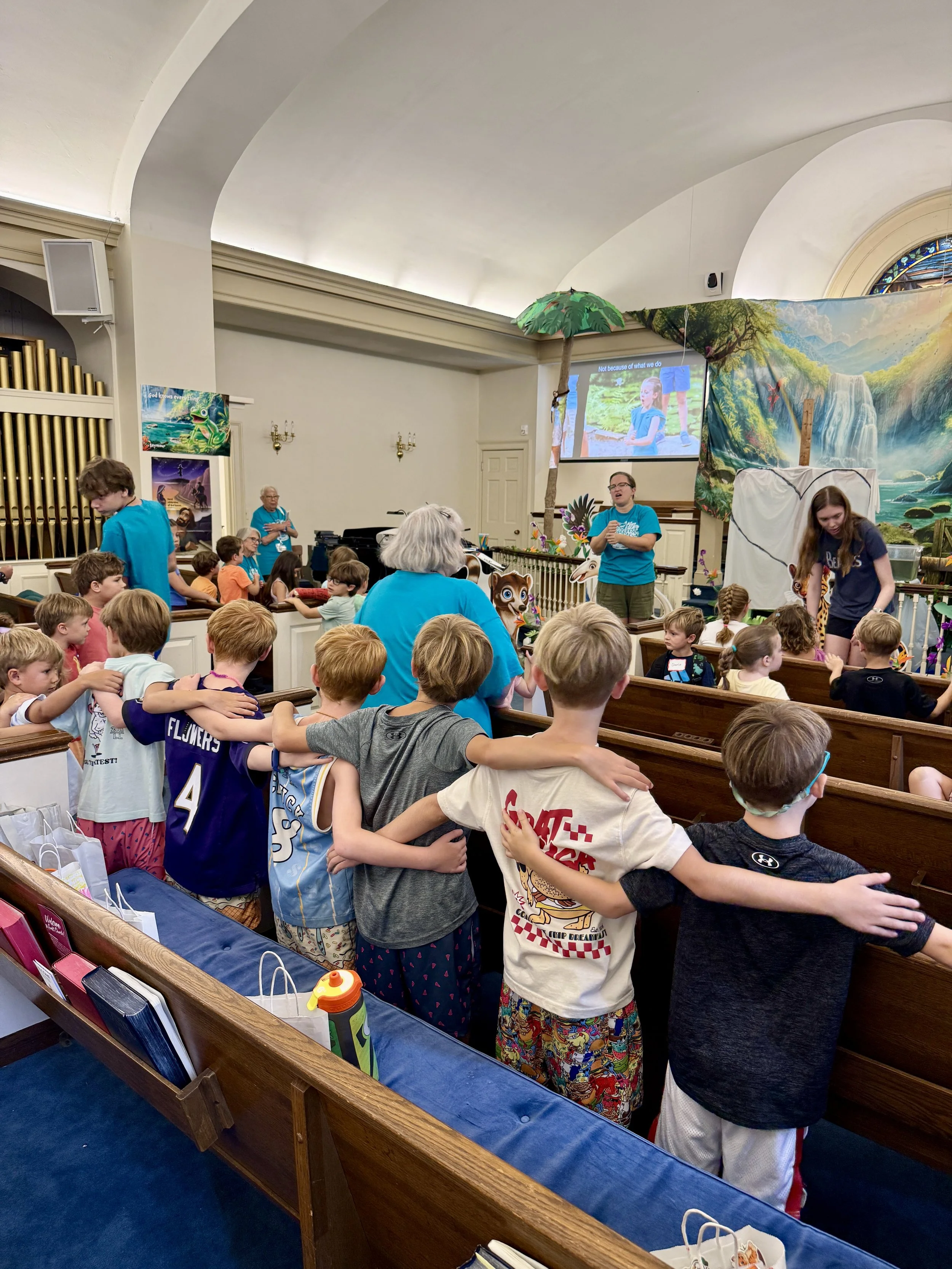 A group of children and adults gathered inside a church, listening to a presentation about animals and nature with a colorful mountain landscape backdrop, decorated with jungle-themed props, including a large tree and animal cutouts.
