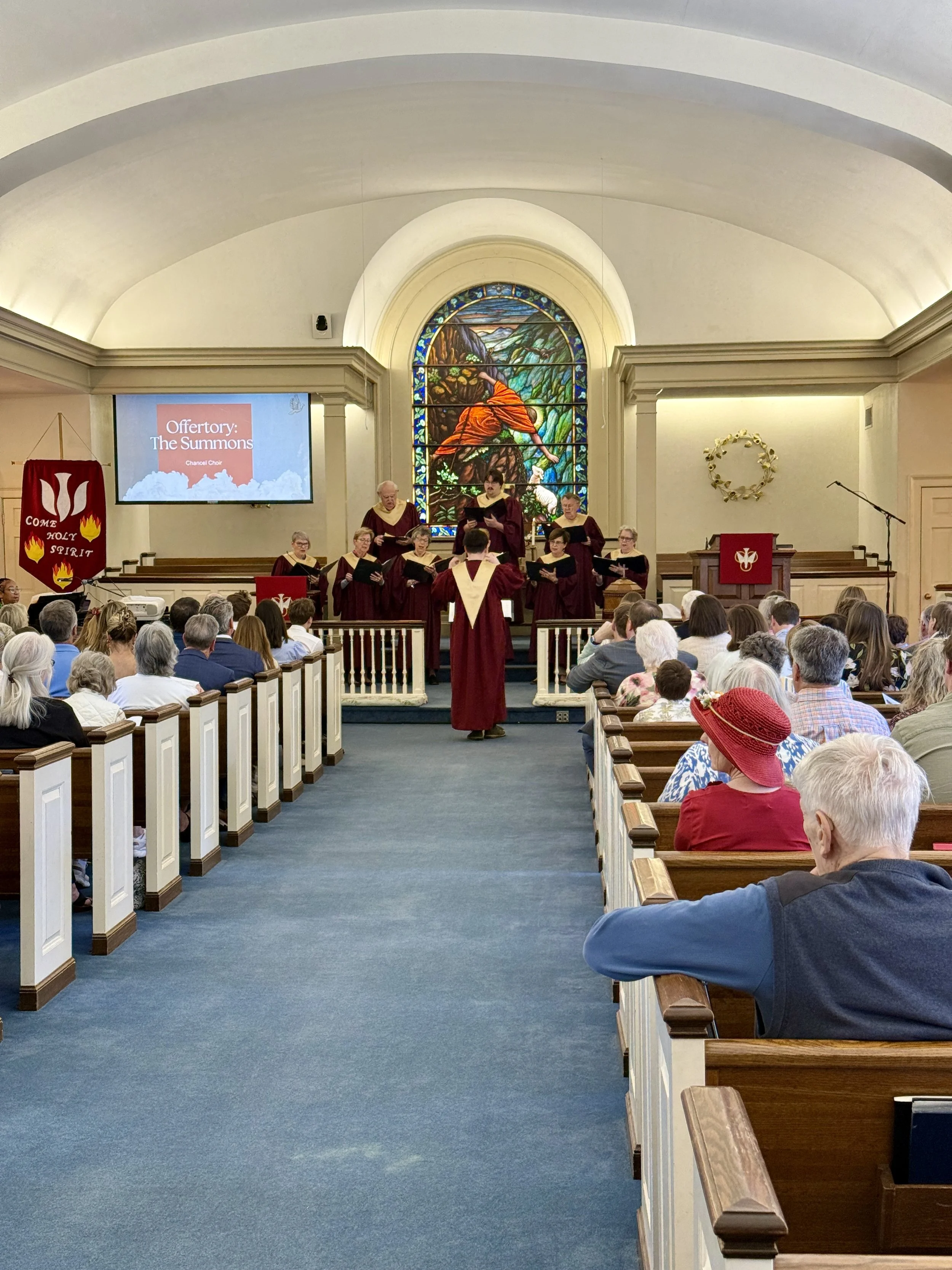 A choir dressed in maroon robes performing on a church stage with a colorful stained glass window of Jesus and nature behind them. The congregation is seated in wooden pews facing the stage.
