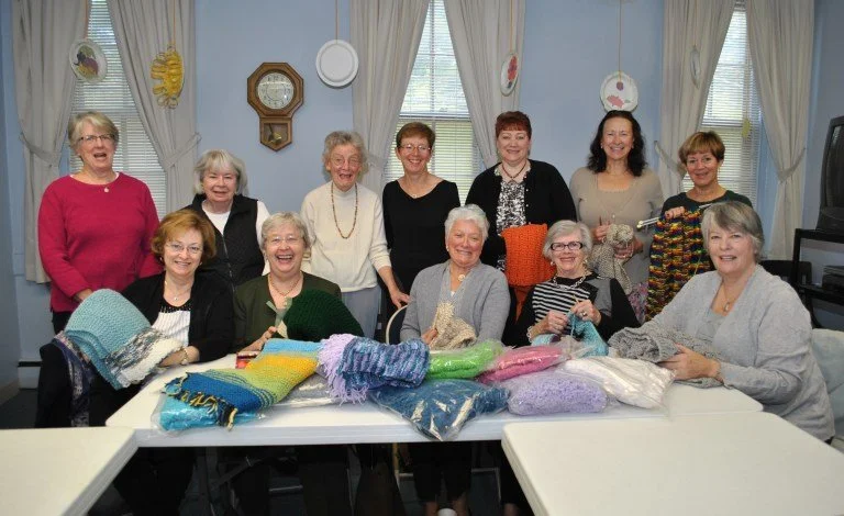 A group of ten women, some holding knitting needles and yarn, gathered around tables with colorful yarns in a bright room with curtains and decorative plates on the wall.