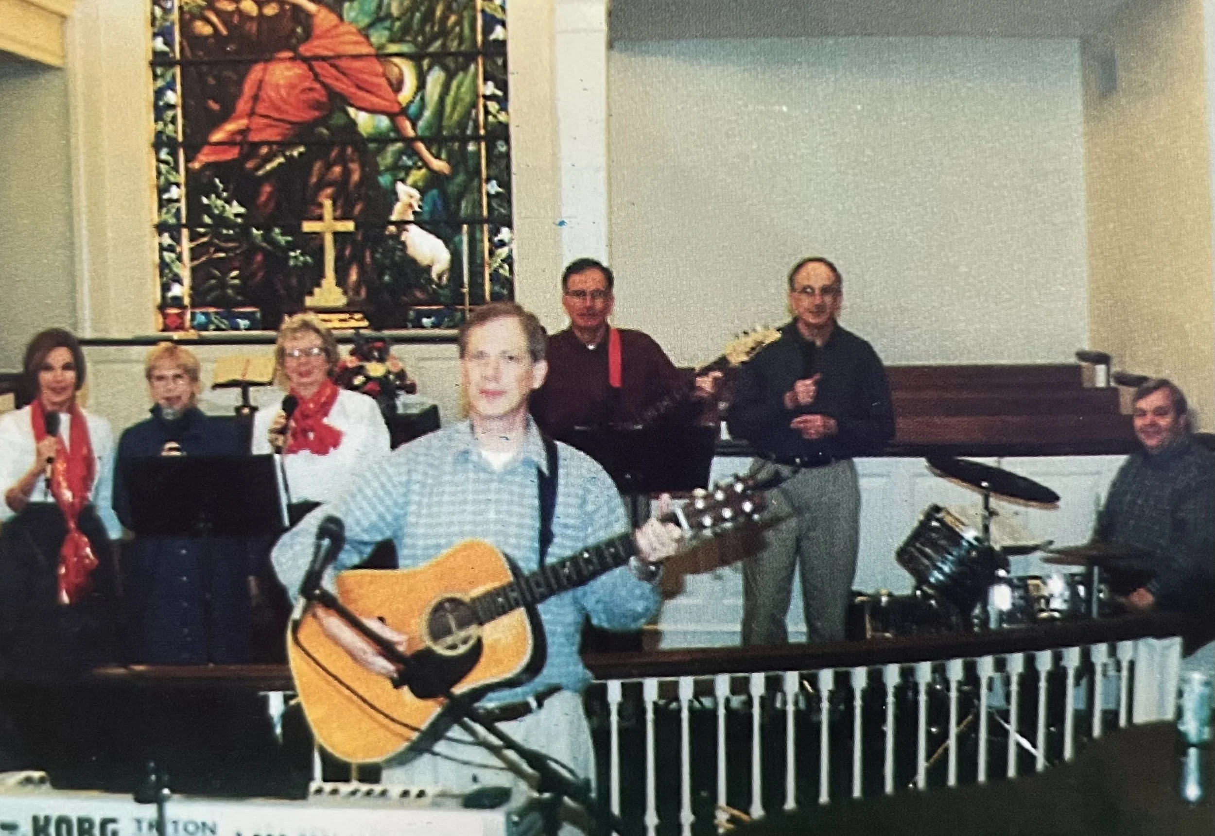 A group of people, including men and women, in a church or religious setting. One person is playing an acoustic guitar, and another is holding a microphone. Some individuals are holding flowers, and a colorful stained glass window featuring a religious figure, a lamb, and a cross is visible in the background. The scene suggests a religious or musical gathering or celebration.