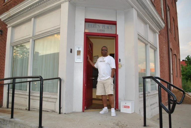 Man standing at the entrance of a building named Earl's Place, located at 1404, with a red door and a white exterior, next to large windows, and a ramp on each side for accessibility.