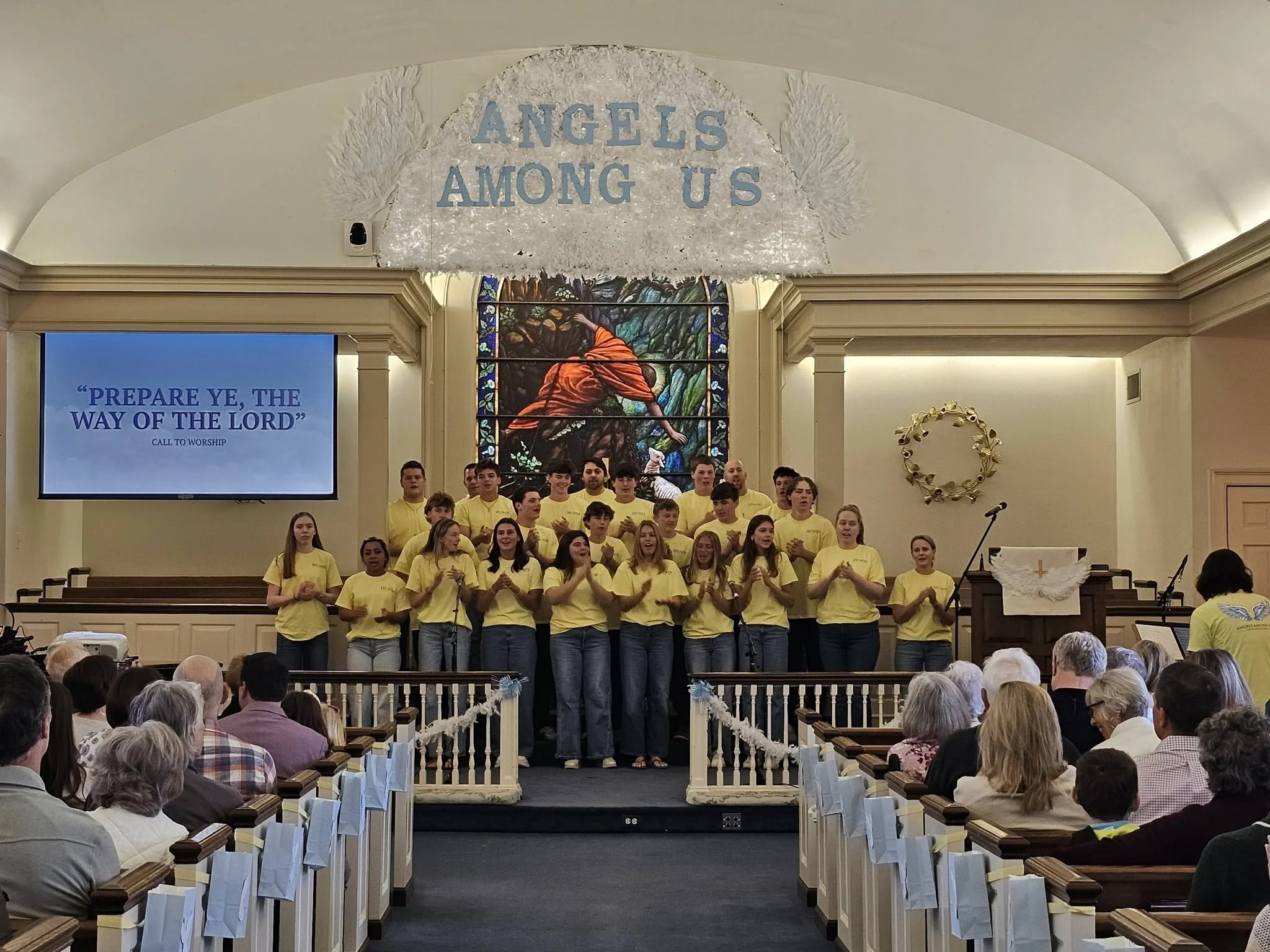 A group of youths in yellow shirts standing on the stage of a church, clapping. Behind them, stained glass window depicting a religious scene. The church is full of seated adults. Ball of flowers or decorations on the wall to the right. A banner above reads 'ANGELS AMONG US'.