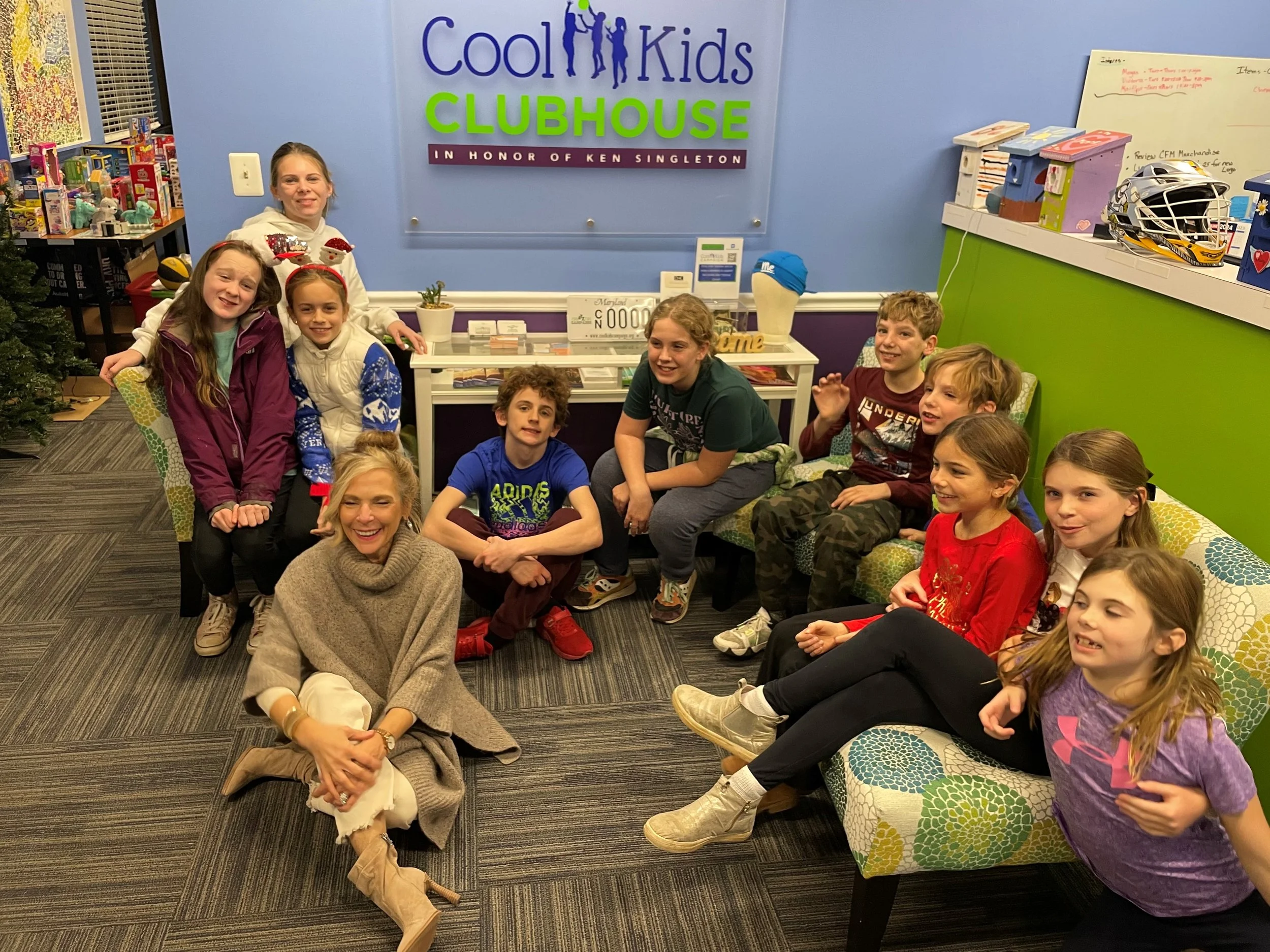 Group of children and an adult woman sitting and kneeling on the floor in a room with a sign that reads 'Cool Kids Clubhouse' on the wall behind them. The children appear happy and are dressed in casual clothes. The room is decorated with colorful furniture and toys, and there is a desk with books and a whiteboard to the right.