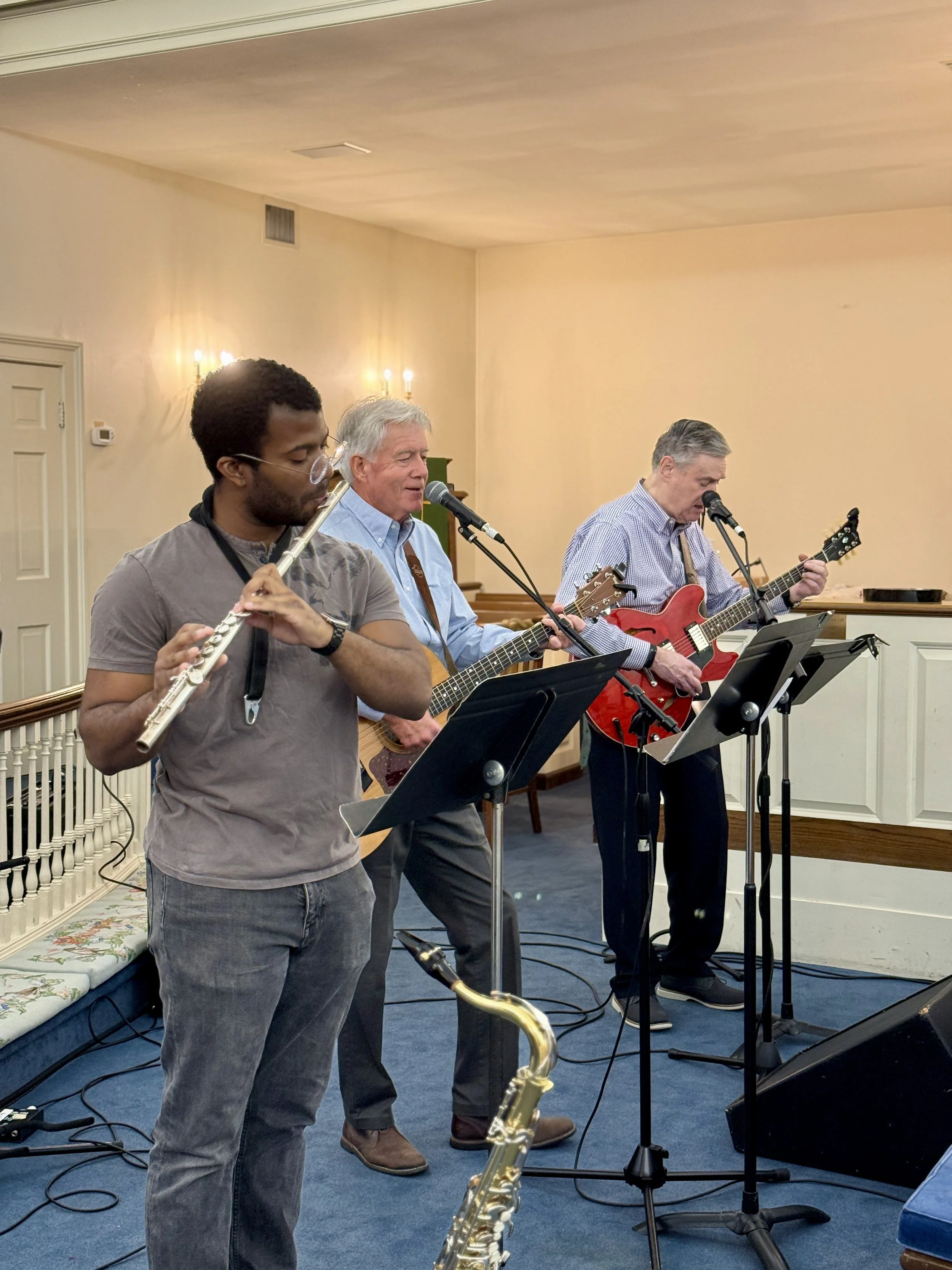 Three men performing music indoors. The man on the left is playing a flute. The two men on the right are singing into microphones, with the man in the middle playing an acoustic guitar and the man on the right playing an electric guitar. All three are standing behind music stands. The room has beige walls, a blue carpet, and wall sconces.