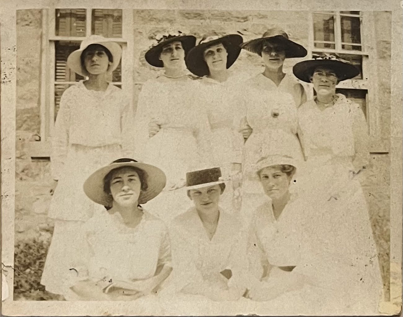 Black and white photo of eight women wearing white dresses and large wide-brimmed hats, standing and sitting outdoors in front of a stone building with windows.