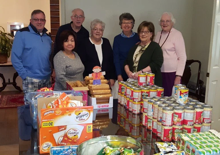 Group of seven people standing behind a table filled with canned goods, boxed food, and snacks in a room with a wooden wall and door.
