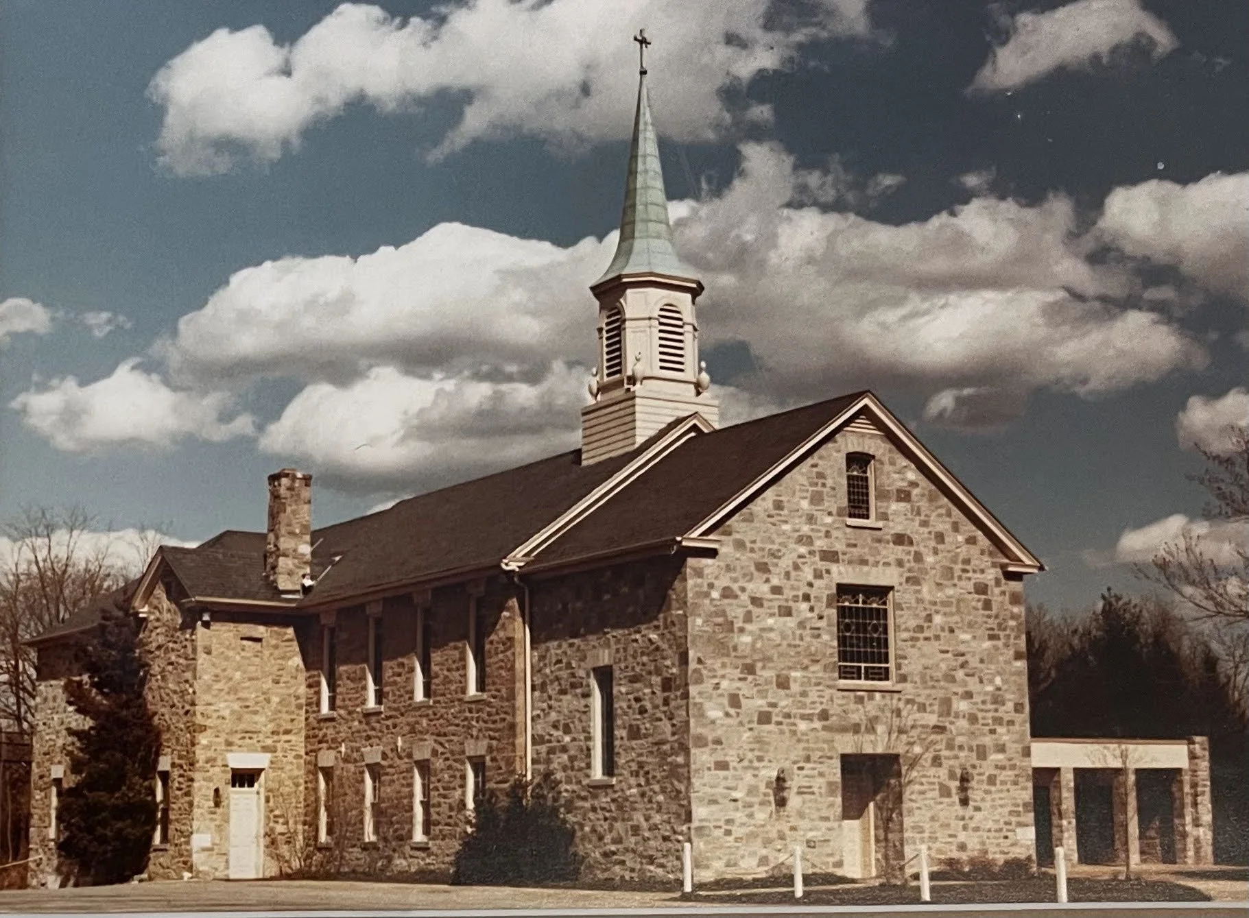 A stone church building with a tall steeple topped by a cross, set against a cloudy sky.