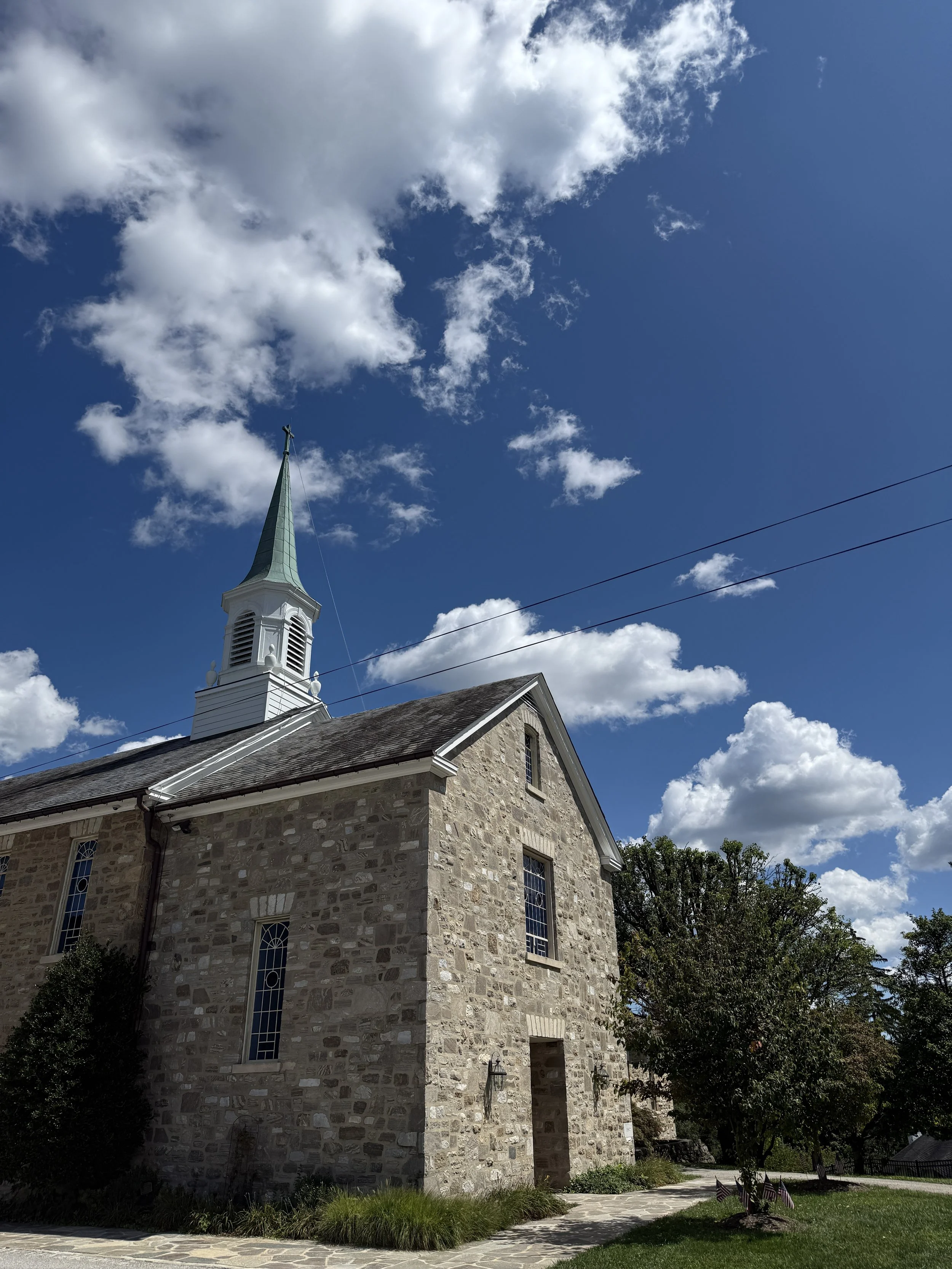 A stone church with a white steeple and green roof, against a partly cloudy blue sky, with small American flags and greenery in front.