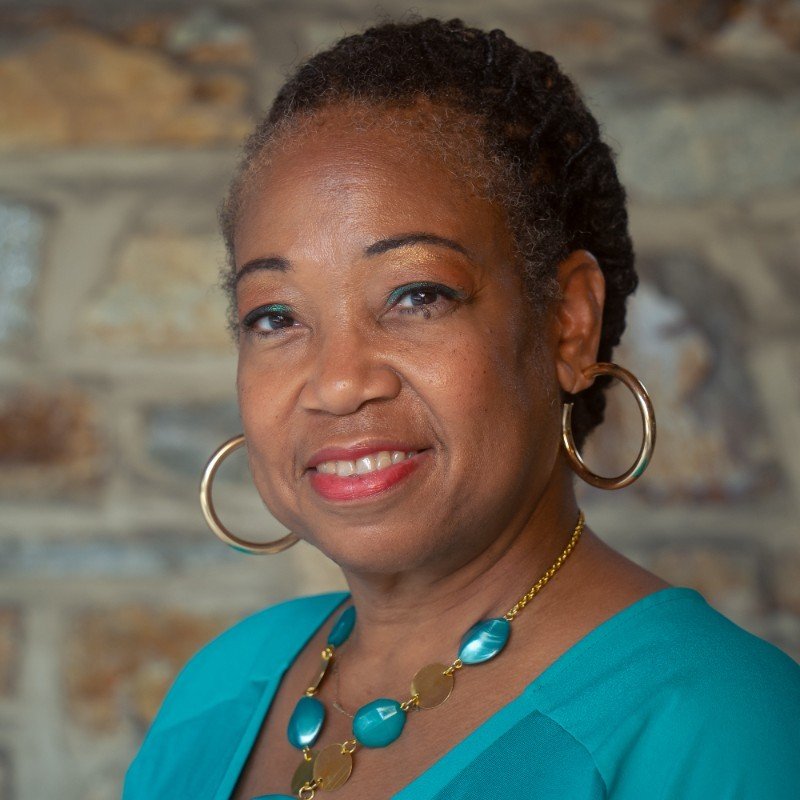 A middle-aged African American woman with short, curly hair styled in twists, wearing large gold hoop earrings, a turquoise necklace with oval beads, and a matching turquoise top, standing in front of a stone wall background.