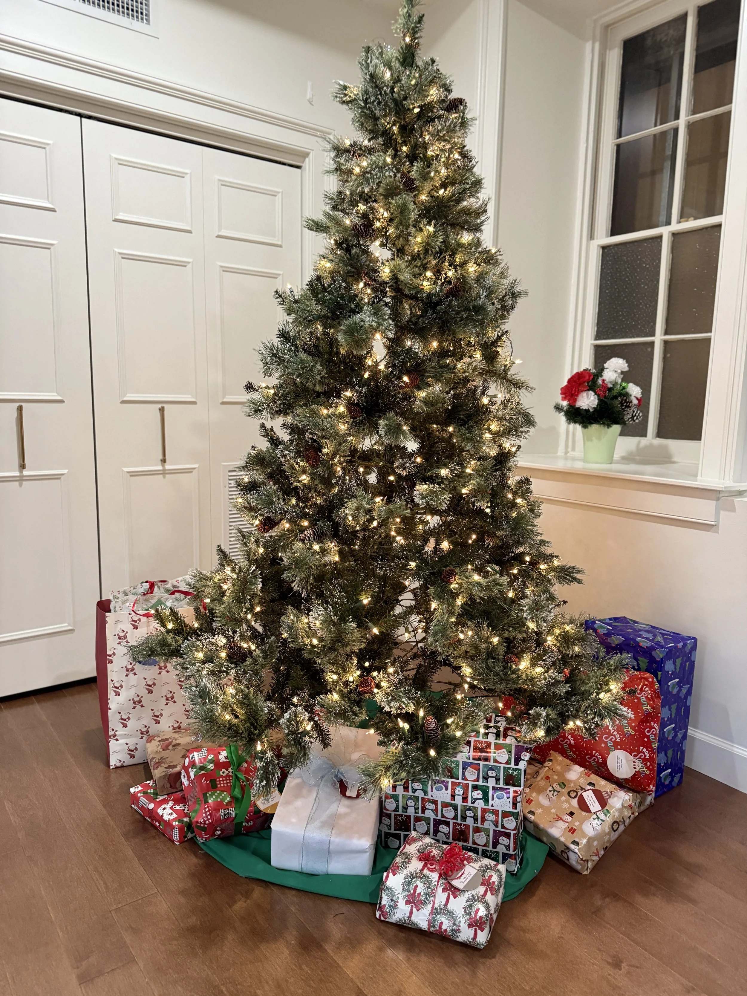 A decorated Christmas tree with white string lights, surrounded by wrapped presents in festive paper on a wooden floor in a room with white walls, a window, and a white door.