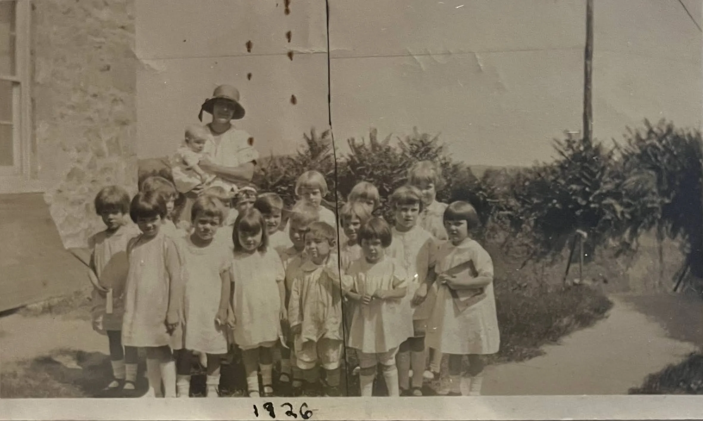 A black and white photo showing a group of children and one adult woman outdoors, with trees and bushes in the background. The children are wearing light-colored dresses or uniforms, and the woman is dressed in light clothing with a hat. The photo has the year '1926' written at the bottom.