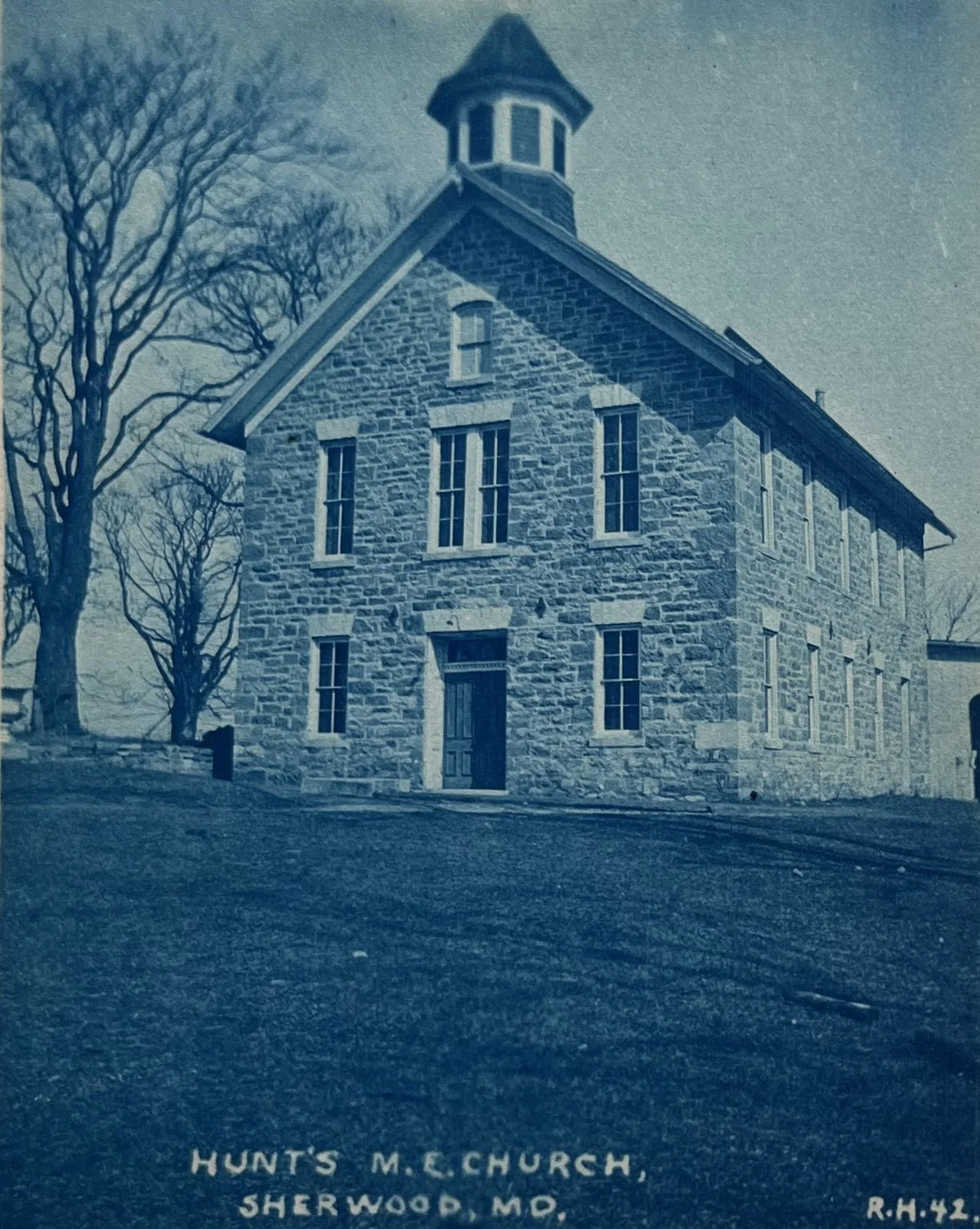 A stone church building with a steeple and tall windows, surrounded by leafless trees. The ground has some text that reads 'HUNT'S M. E. CHURCH SHERWOOD, MD R.H. 44'.