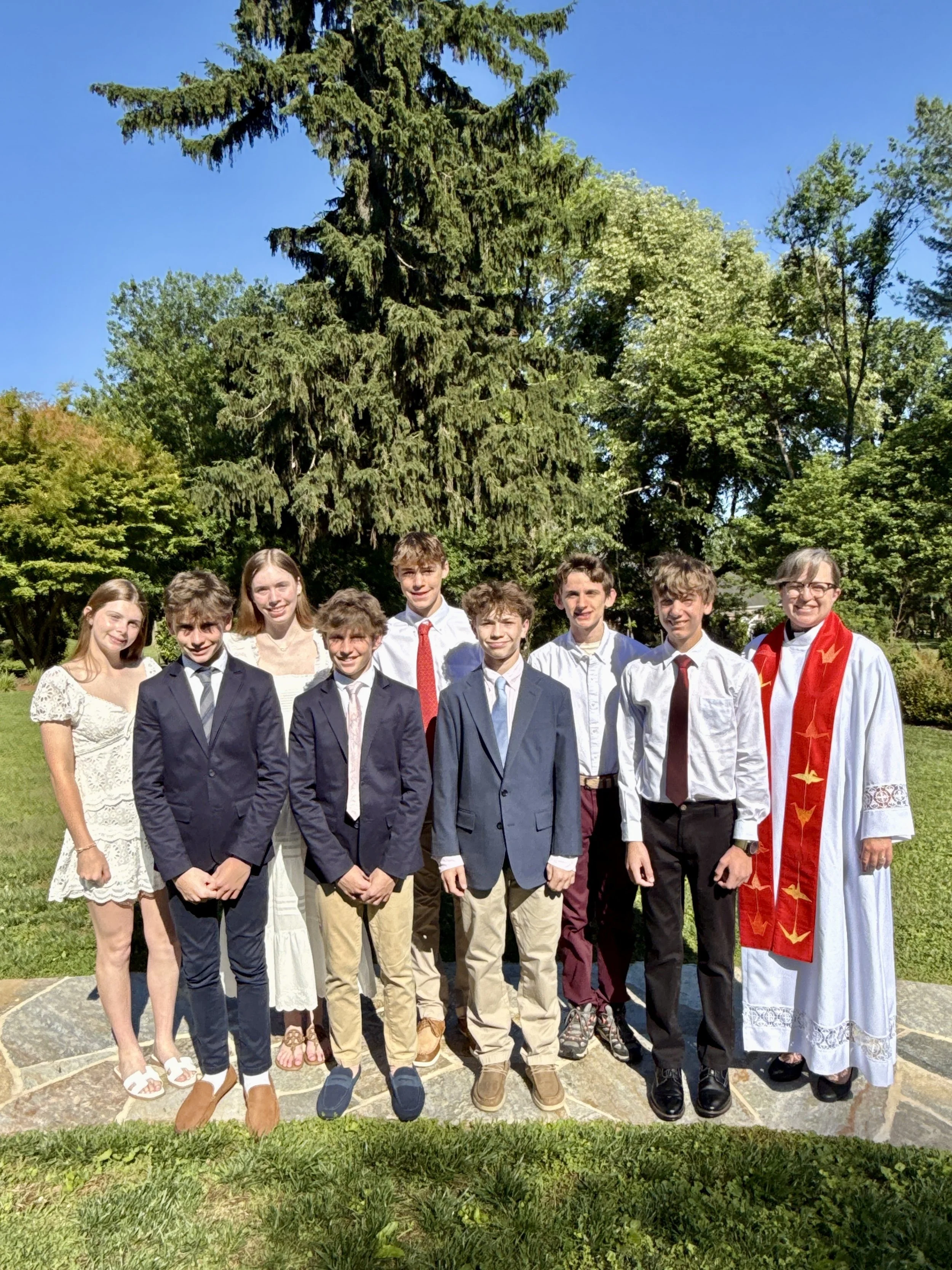 A group of young people standing outdoors on a sunny day in front of green trees, some dressed in formal attire, with one person wearing a white graduation gown and red stole, likely celebrating a graduation.