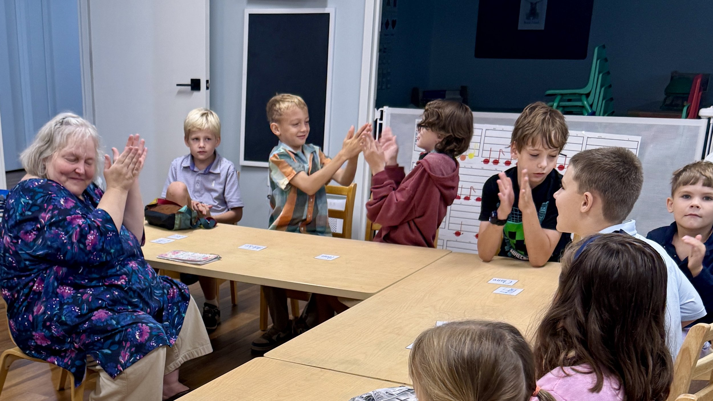 Older woman and children at a table engaging in high-fives and interaction in a classroom setting, with musical notes on a whiteboard in the background.