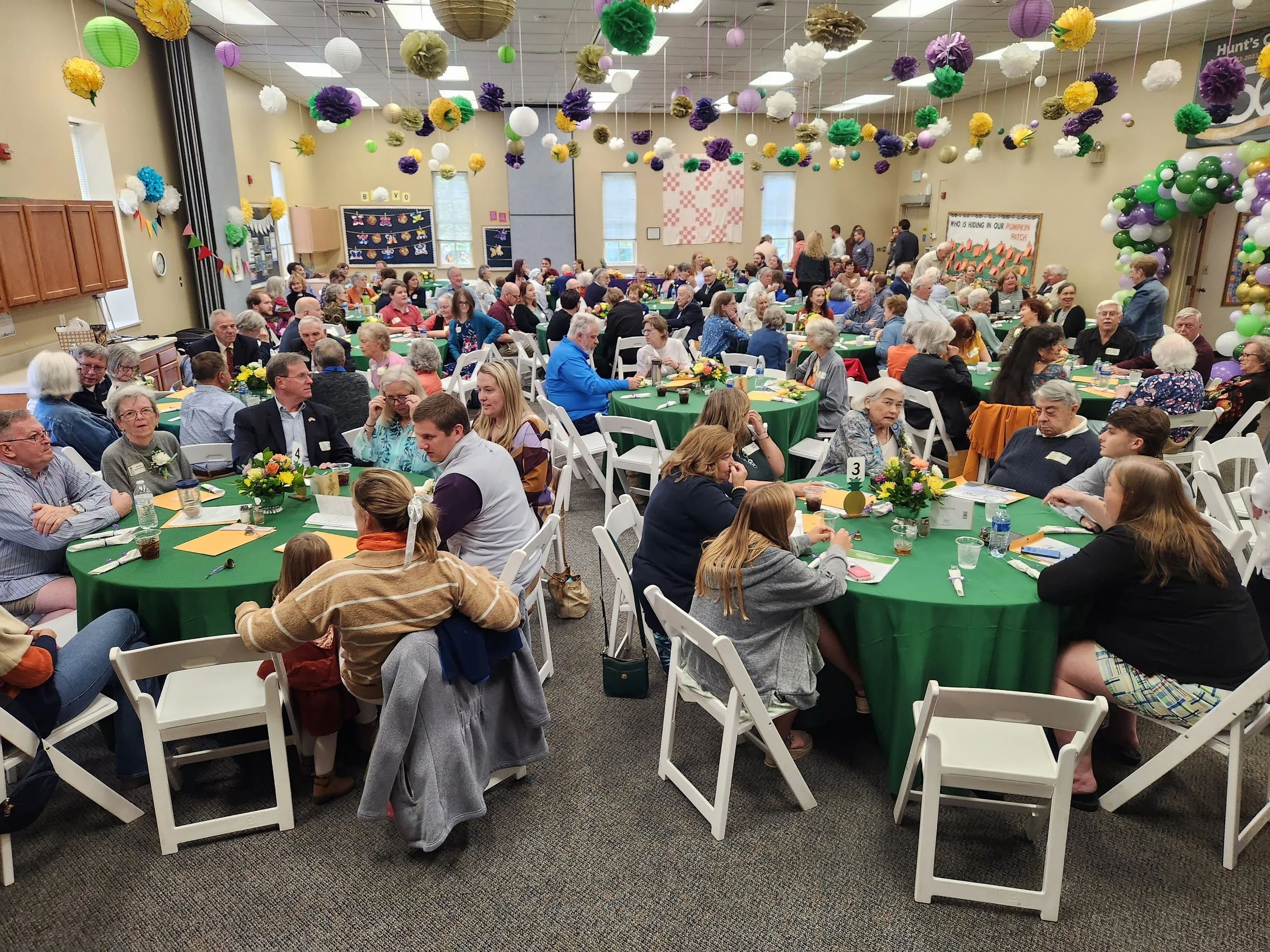 A large room filled with many round tables covered with green tablecloths, each surrounded by white folding chairs. The tables are decorated with colorful flower arrangements. The room is decorated with hanging paper lanterns and pom-poms in various colors and sizes on the ceiling. Many people of various ages are sitting at the tables, eating and talking, with some standing and mingling in the background. There are bulletin boards and artwork on the walls, and a balloon arch on one side of the room.