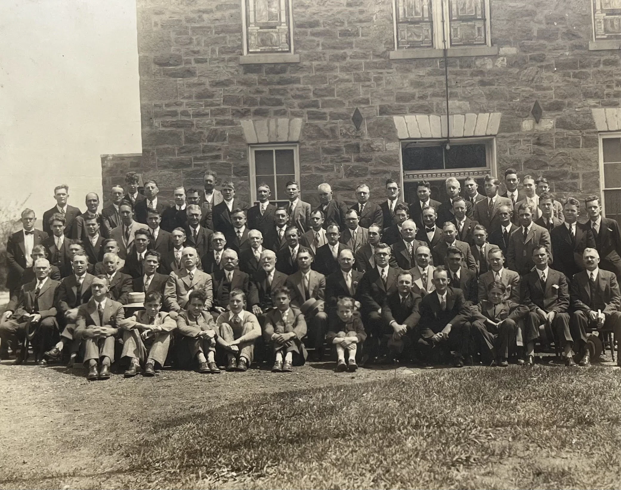 A large group of men dressed in formal suits and ties gathered for a photo in front of a brick building with three windows.