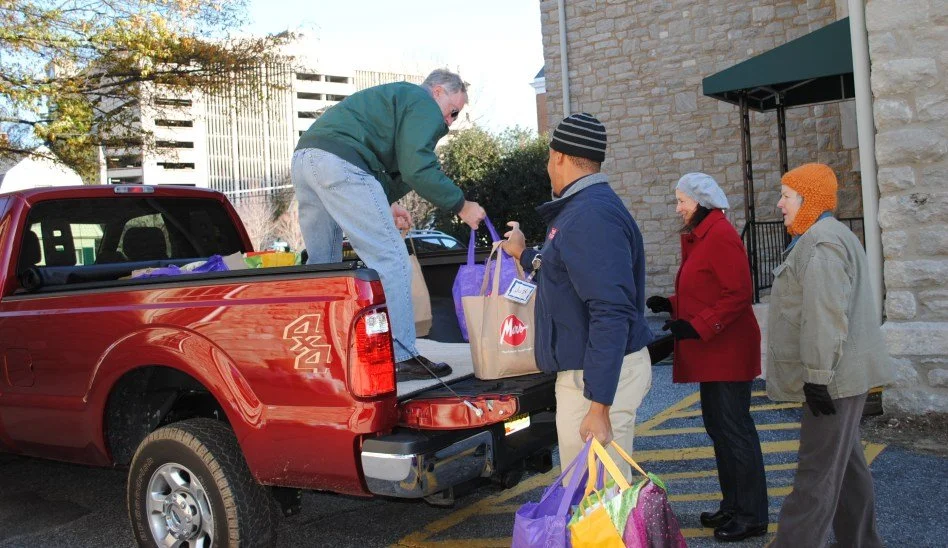 Four people load groceries into the back of a red pickup truck outside a stone building. Two women and one man are waiting in line, all wearing warm clothing and hats.