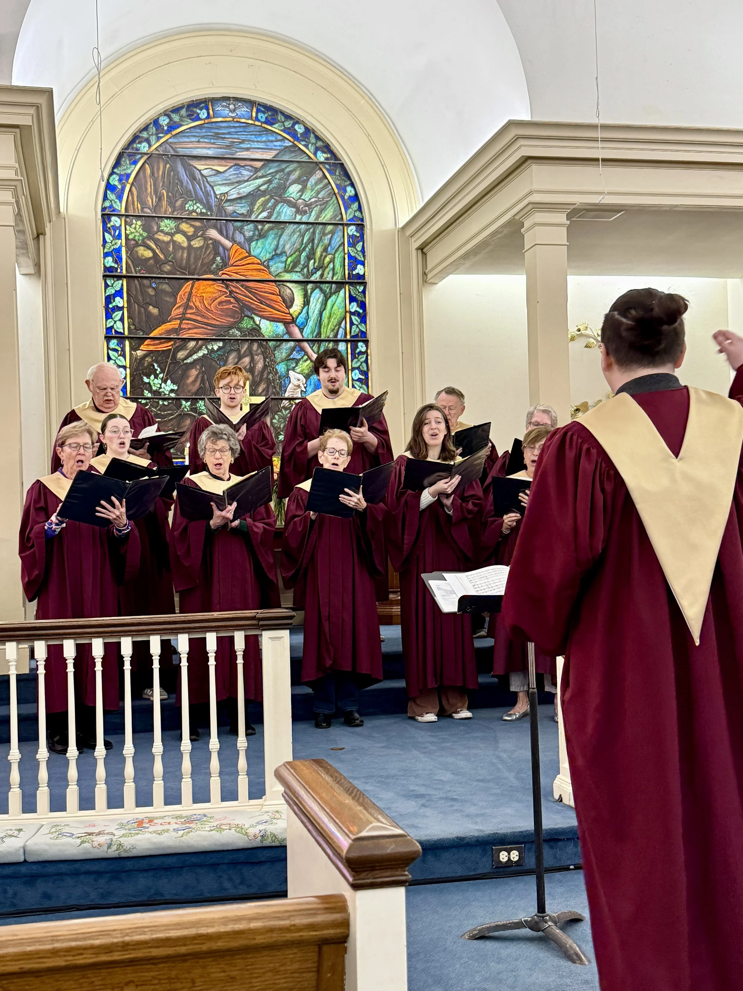 Choir of men and women singing in a church with stained glass window showing a biblical scene.