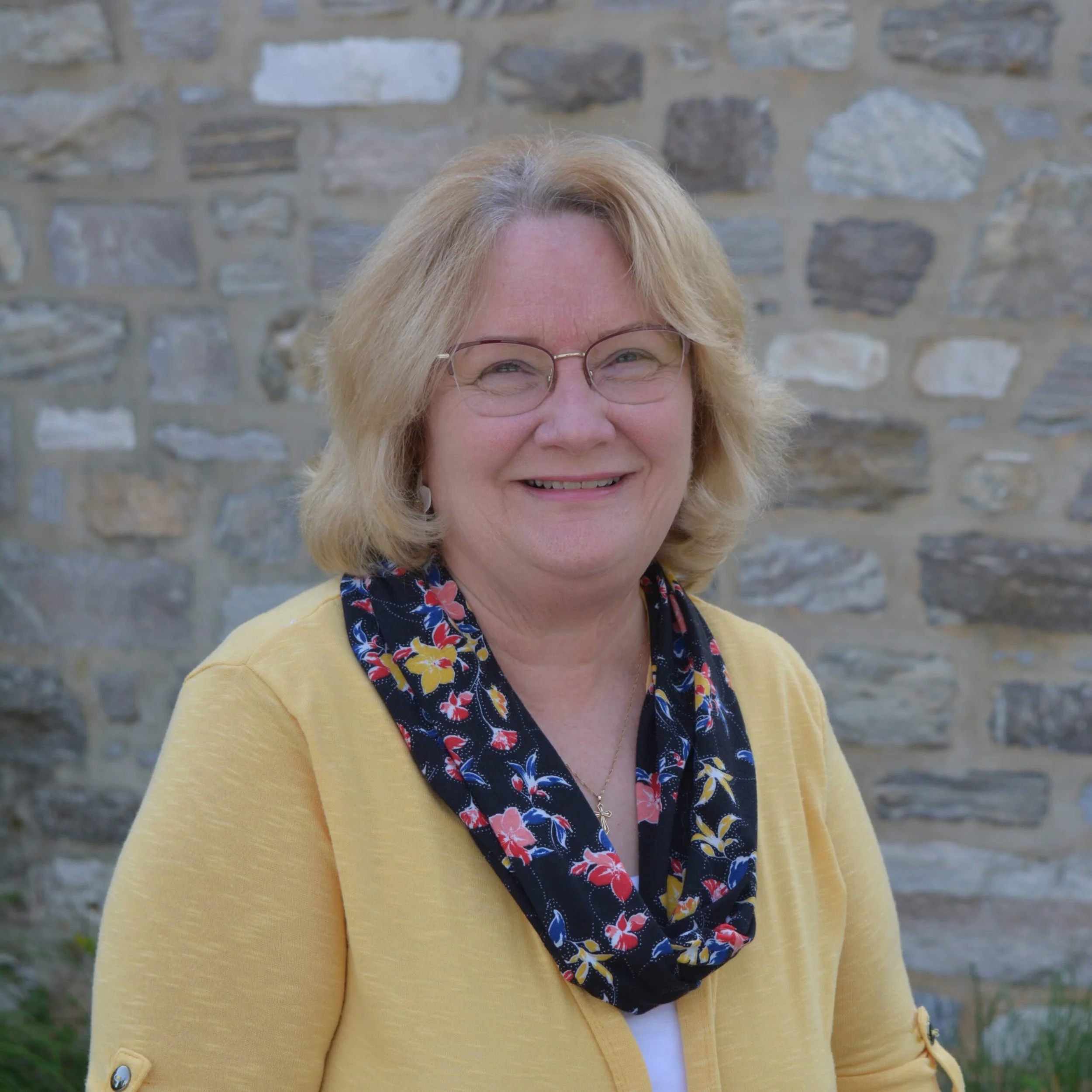 Close-up of a smiling woman with blonde hair, wearing glasses, a yellow cardigan, a floral blouse, and a necklace, standing outdoors in front of a stone wall.