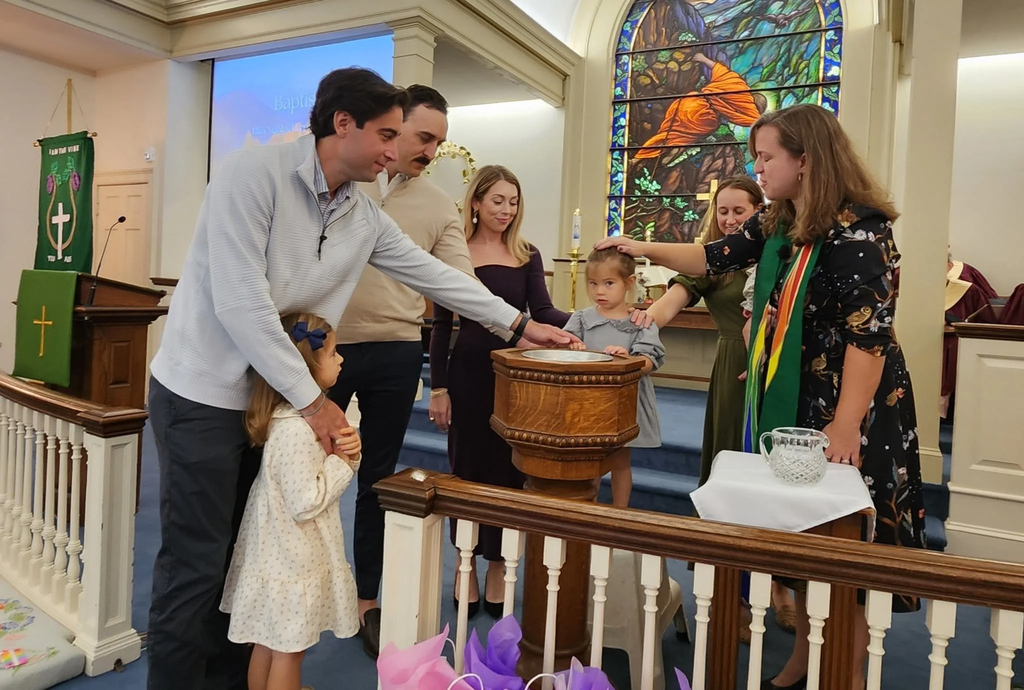 A baptism ceremony in a church with children and adults gathered around a baptismal font, participating in a religious ritual.