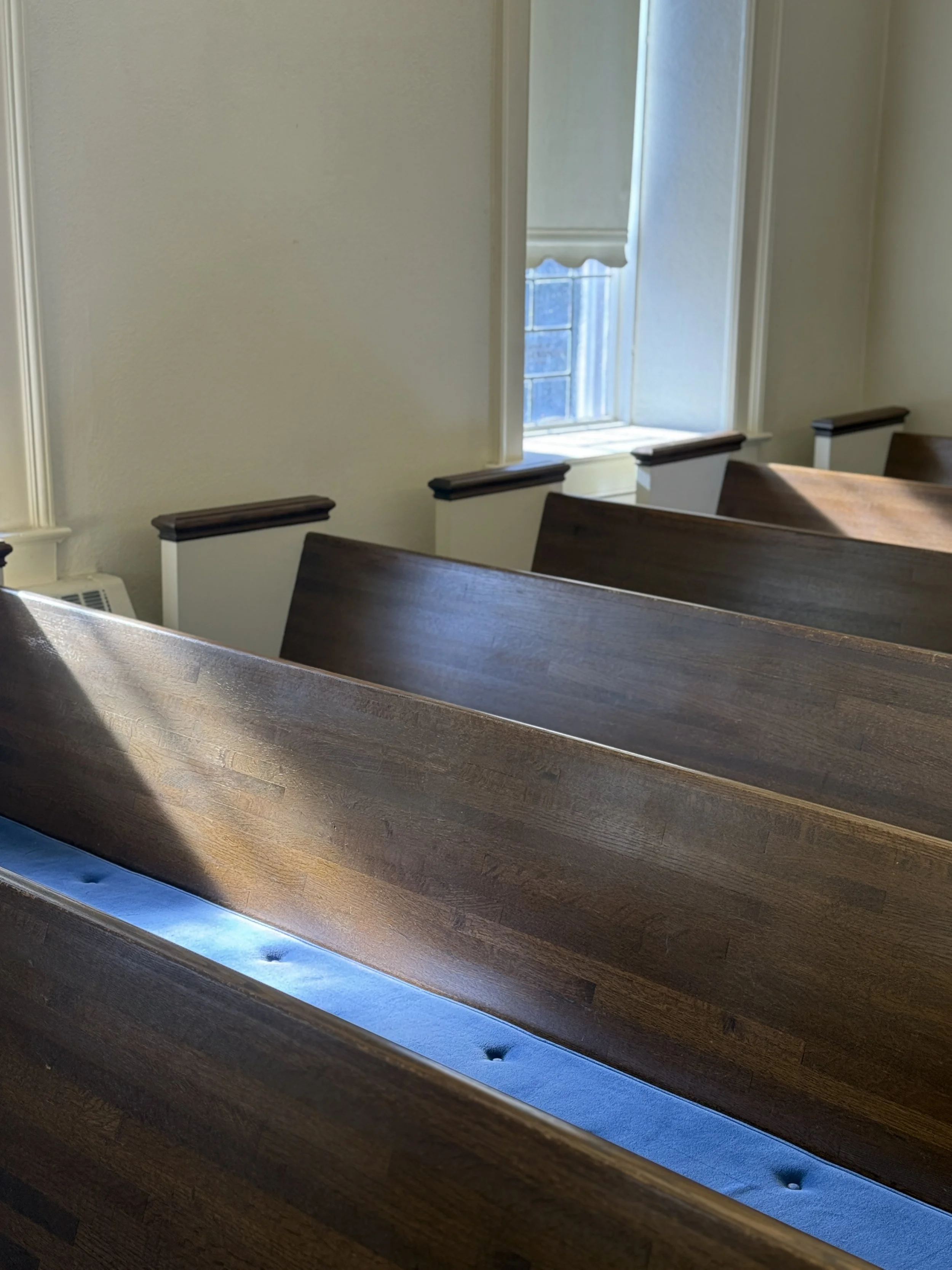 View of wooden church pews with a blue light at the base, overlooking a window with sunlight shining through.