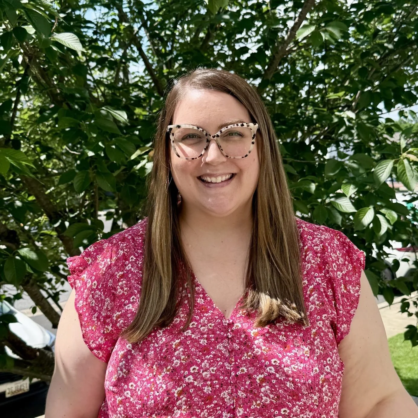 A woman with long brown hair, wearing large tortoiseshell glasses and a pink floral dress, smiling outdoors in front of green leafy trees.