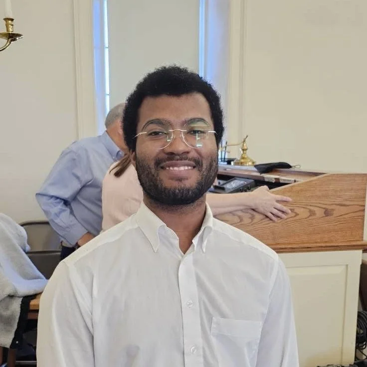 A smiling man with glasses and a beard wearing a white dress shirt standing inside a room with cream-colored walls and large windows.