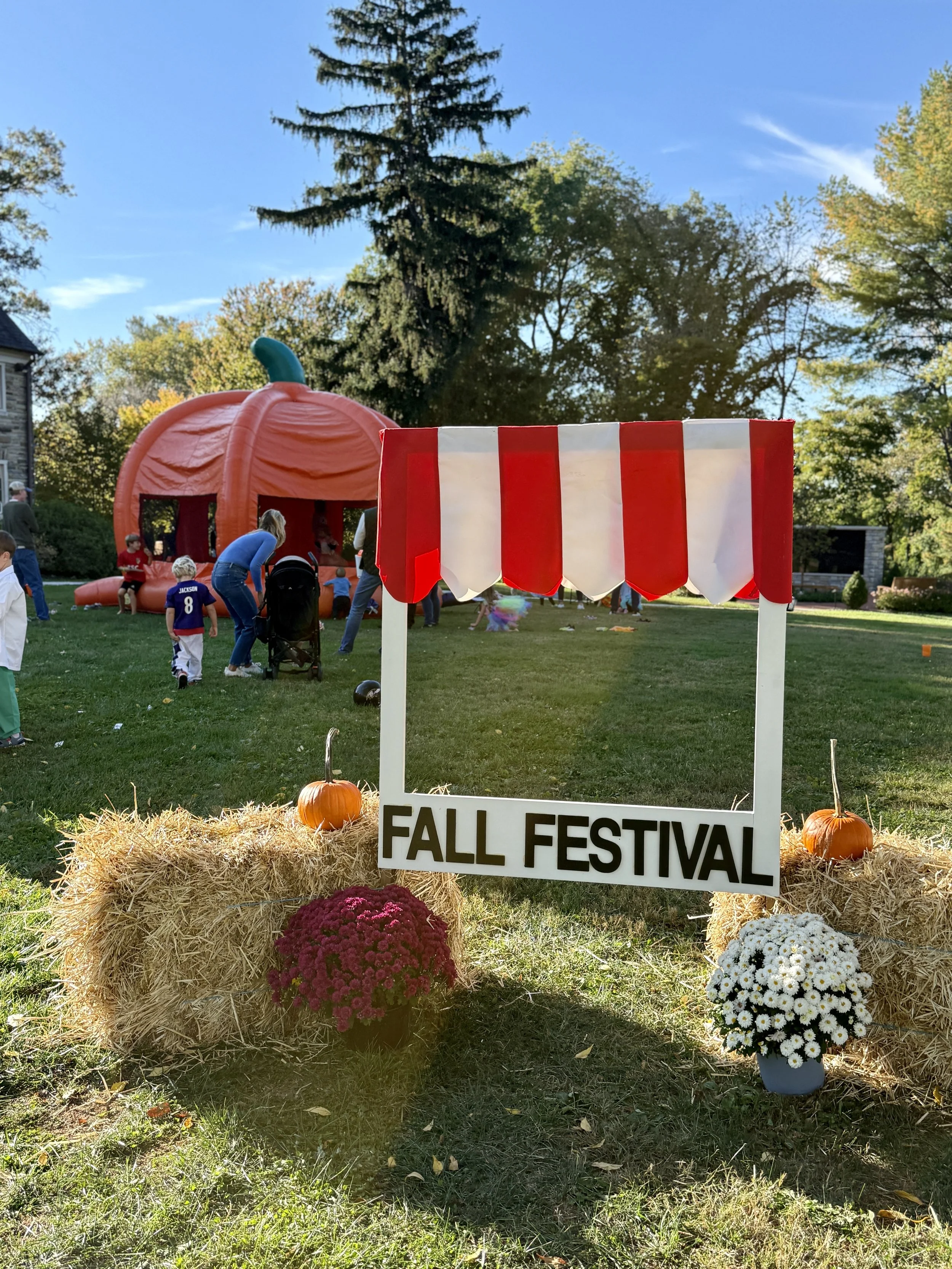 Decorative fall festival scene with a sign reading 'Fall Festival' in front of hay bales, pumpkins, and colorful chrysanthemums, with a pumpkin-shaped bounce house and people enjoying the event on a sunny day with trees in the background.