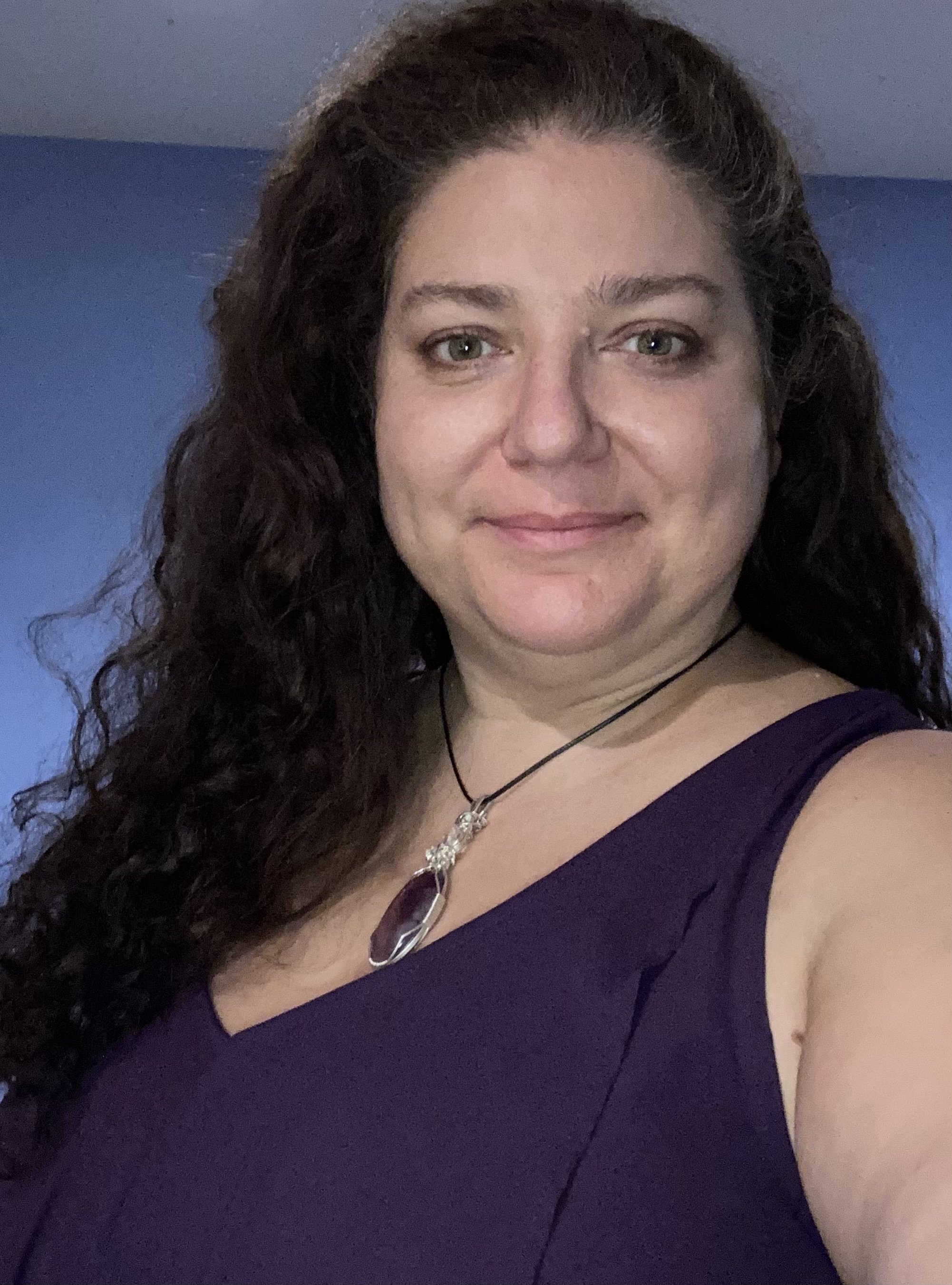 Founder , long curly brown hair, wearing a purple dress and a necklace, smiling.