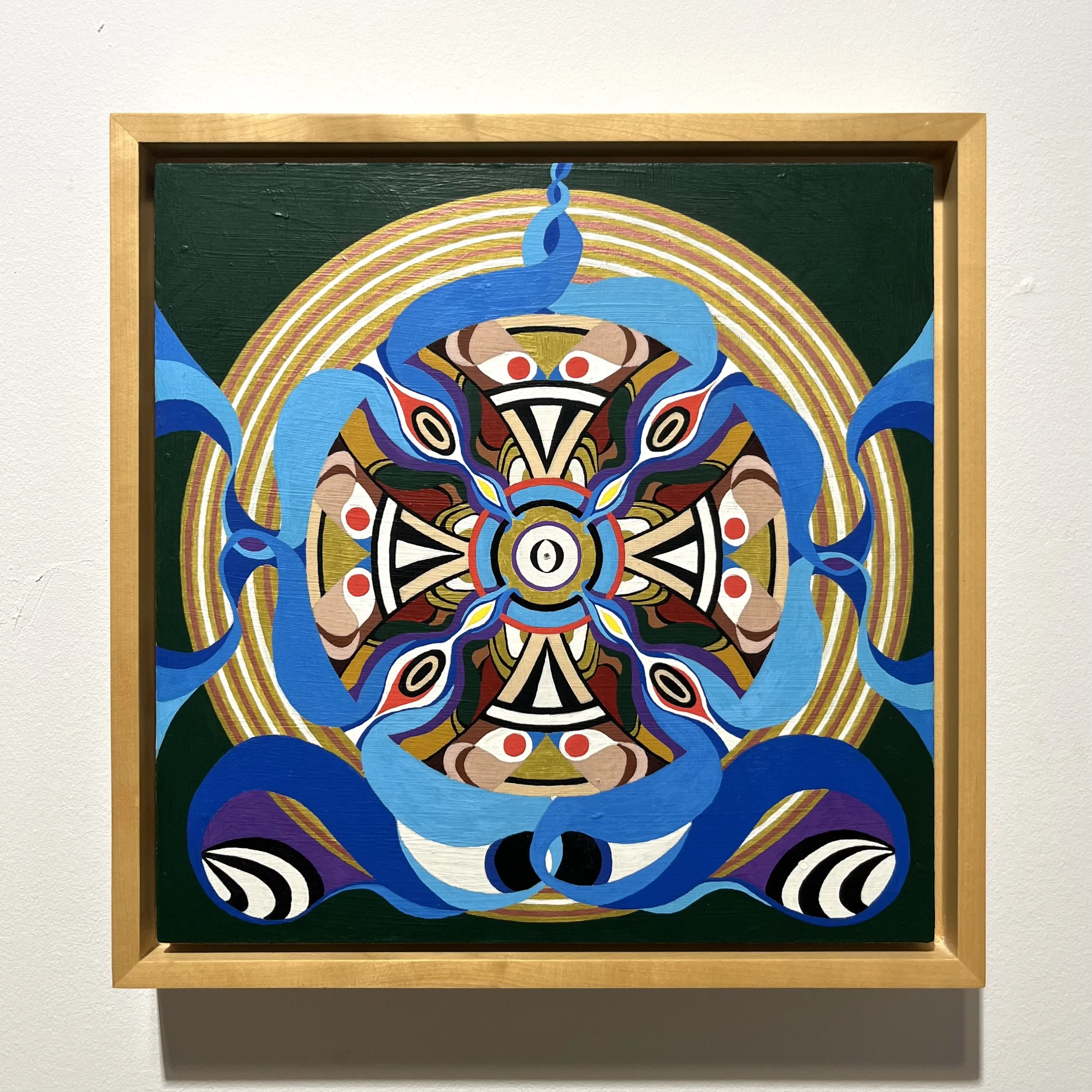 A colorful, abstract painting featuring a symmetrical design with vivid blue, red, yellow, black, white, and brown colors, surrounded by concentric rainbow-like arcs, housed in a light wooden frame mounted on a white wall.