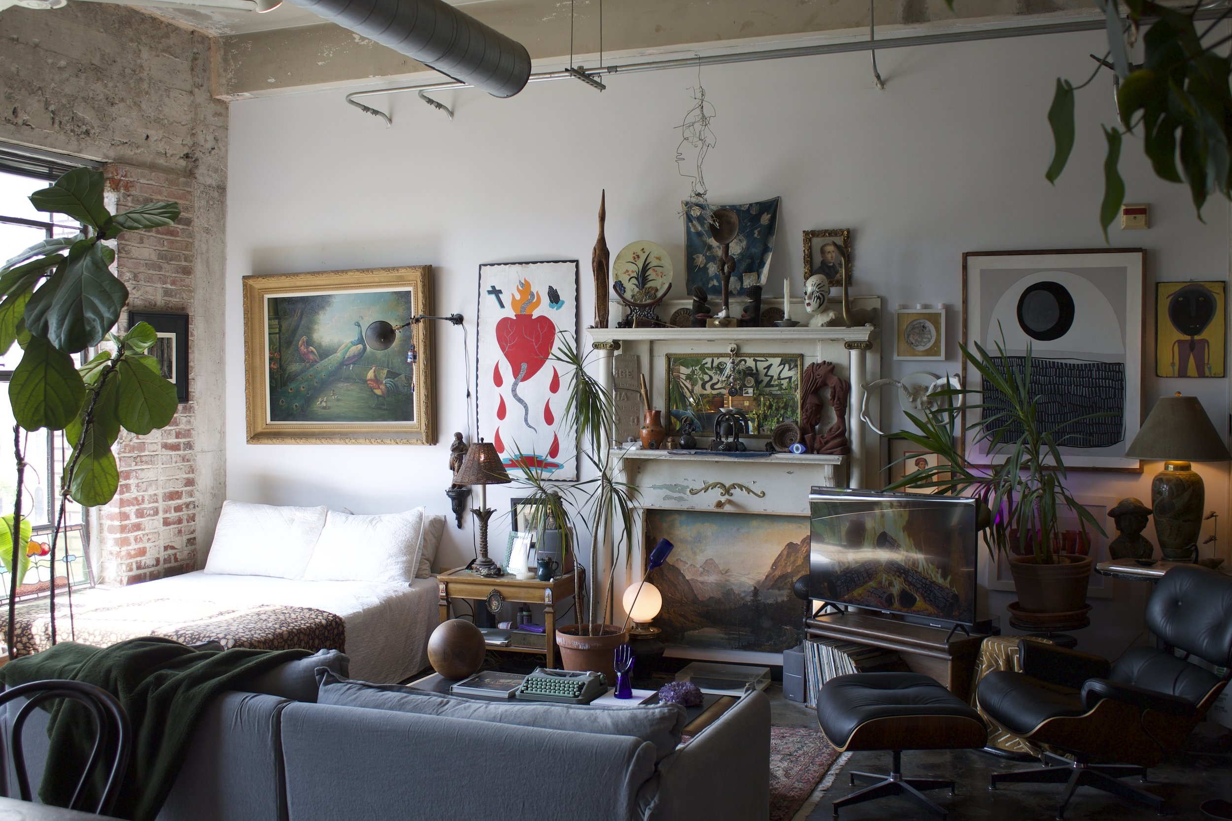 Living room with various art pieces and plants, exposed brick and concrete walls, a mix of modern and vintage furniture, a television, and decorative objects.