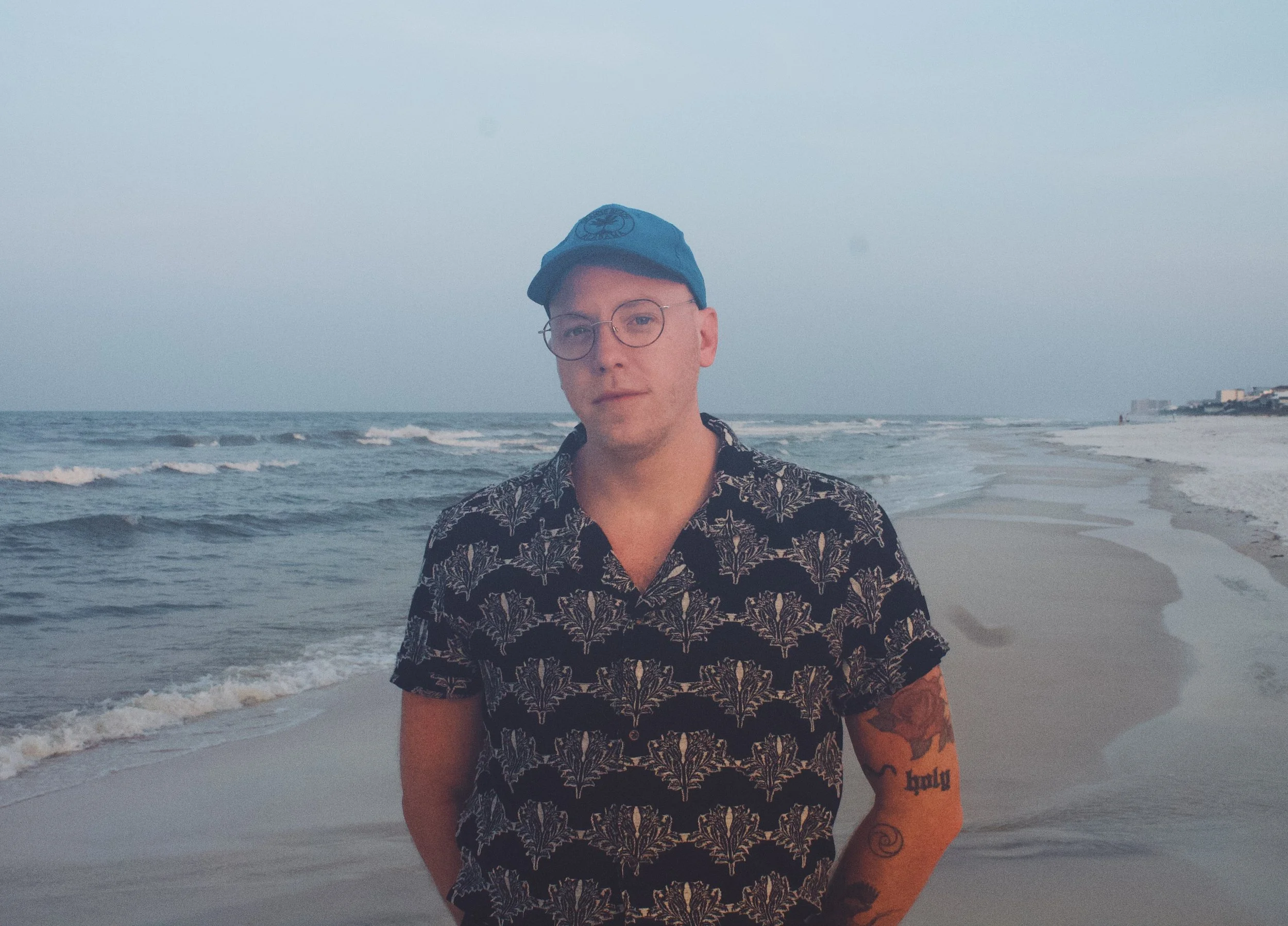 A young man standing on a beach with the ocean and a cloudy sky in the background. He is wearing glasses, a blue cap, and a black shirt with a white floral pattern. He has tattoos on his arms and is looking directly at the camera.
