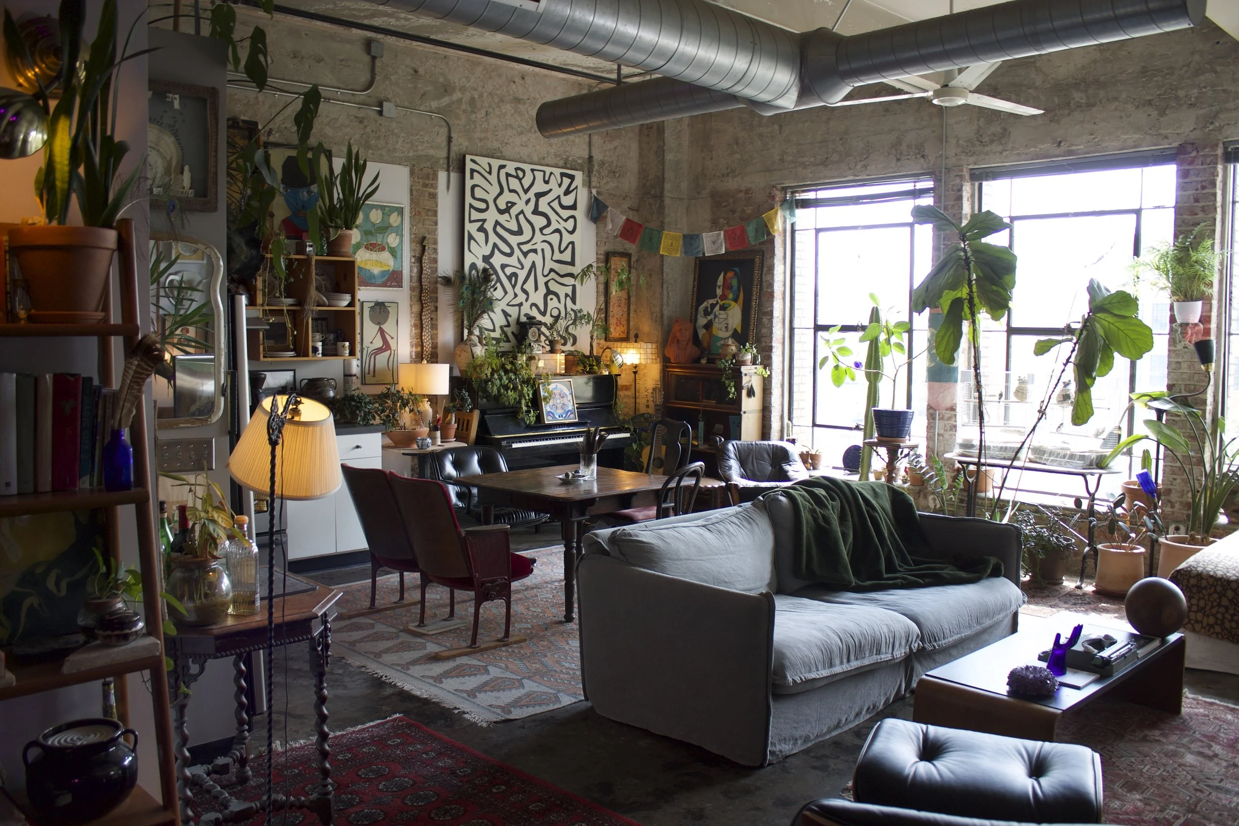 Interior of a cozy urban apartment with exposed brick walls, large windows, plenty of houseplants, a black upright piano with artwork, a wooden dining table with chairs, a gray sofa, and eclectic decor.