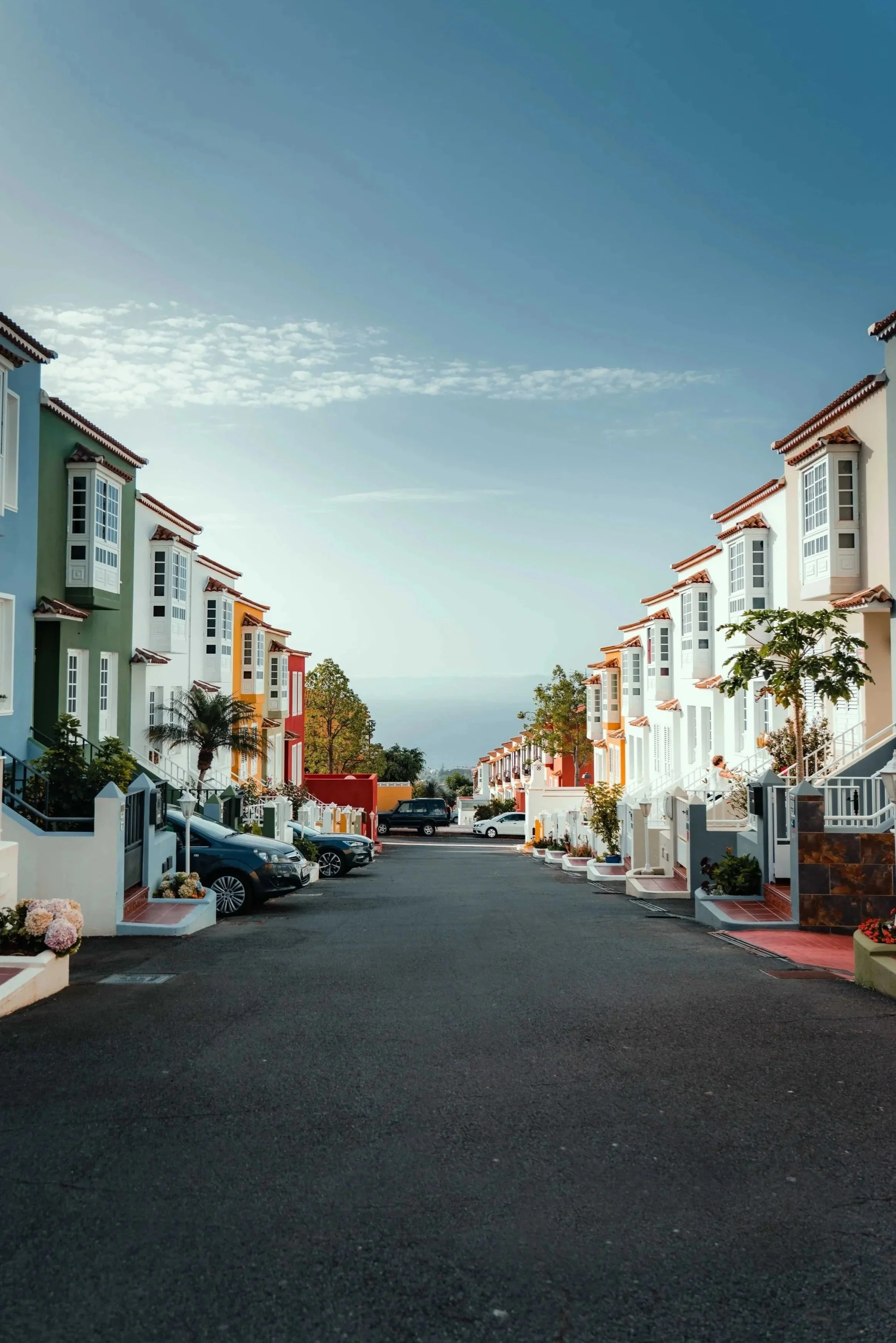 View of a residential street flanked by colorful multi-story houses with red-tiled roofs, parked cars, small trees, and a clear blue sky with wispy clouds.