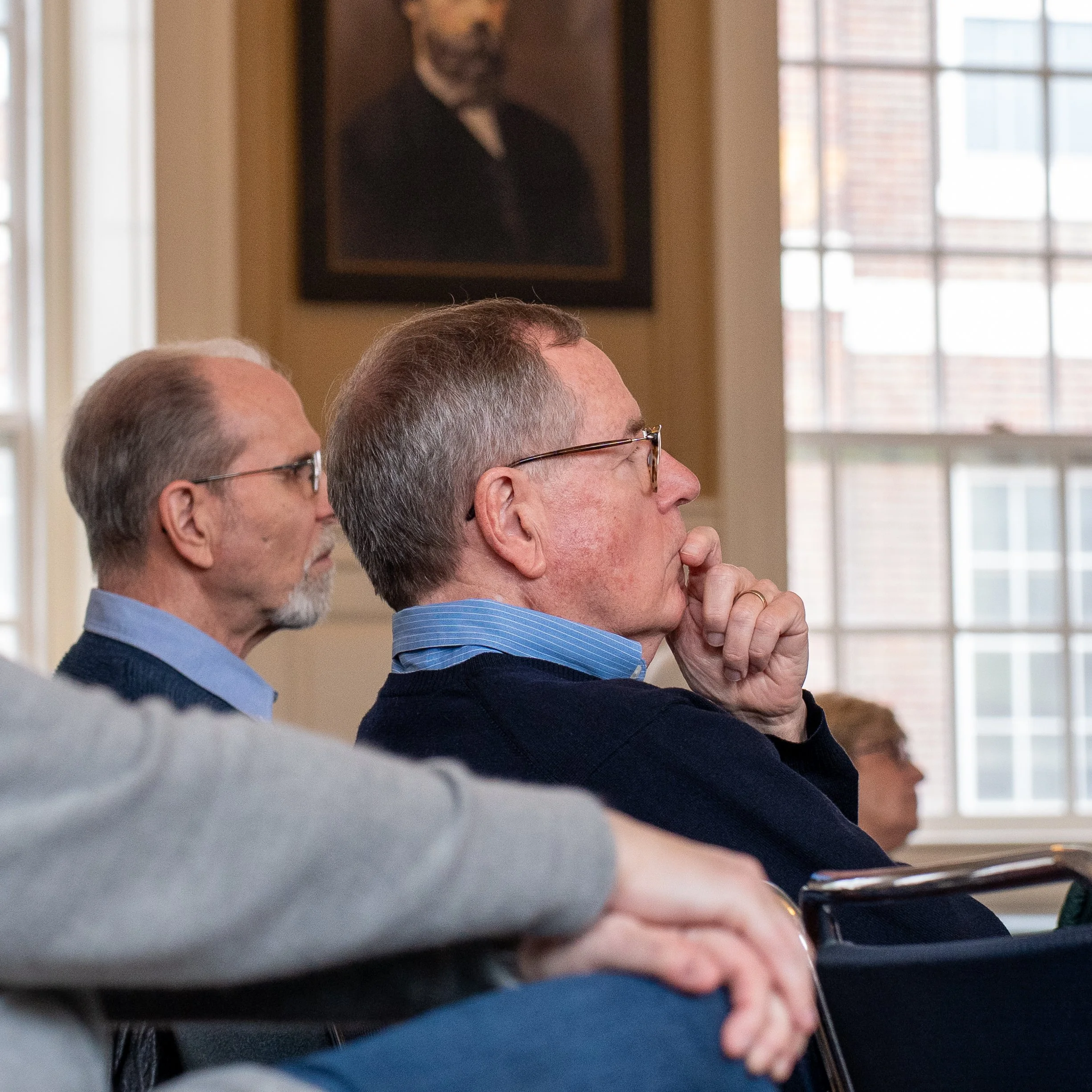 Side profile of older man with glasses and graying hair, possibly participating in a group event, with other people visible in the background, and a portrait hanging on the wall near a window.
