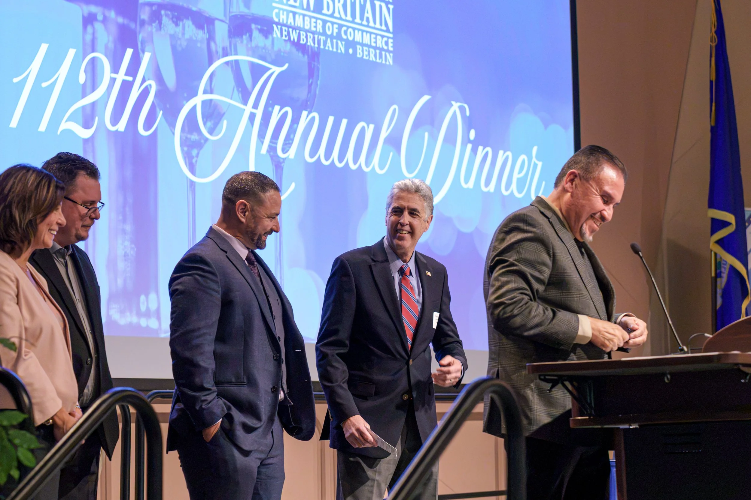 A group of five people on stage at an event celebrating the 120th anniversary of the New Britain Chamber of Commerce, with a large screen behind them displaying the event details. They are smiling and dressed in formal attire.