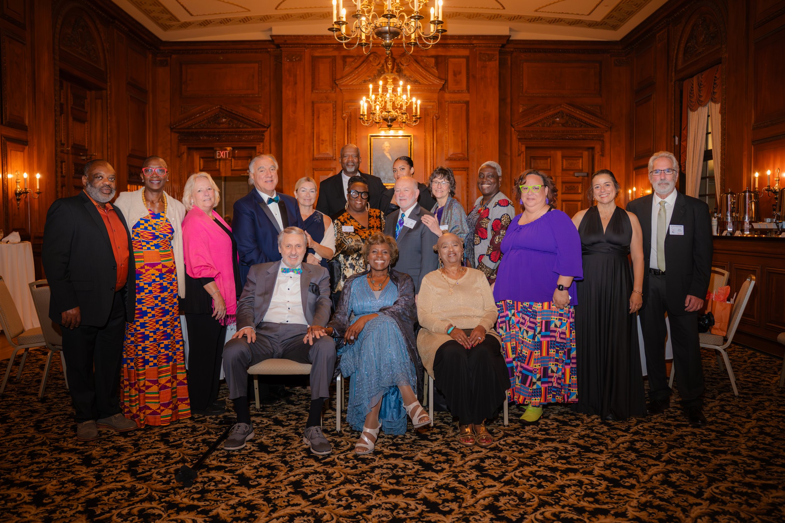 A diverse group of people dressed in formal and colorful attire posing for a photo in an elegant wood-paneled banquet room with chandeliers and a framed portrait on the wall.
