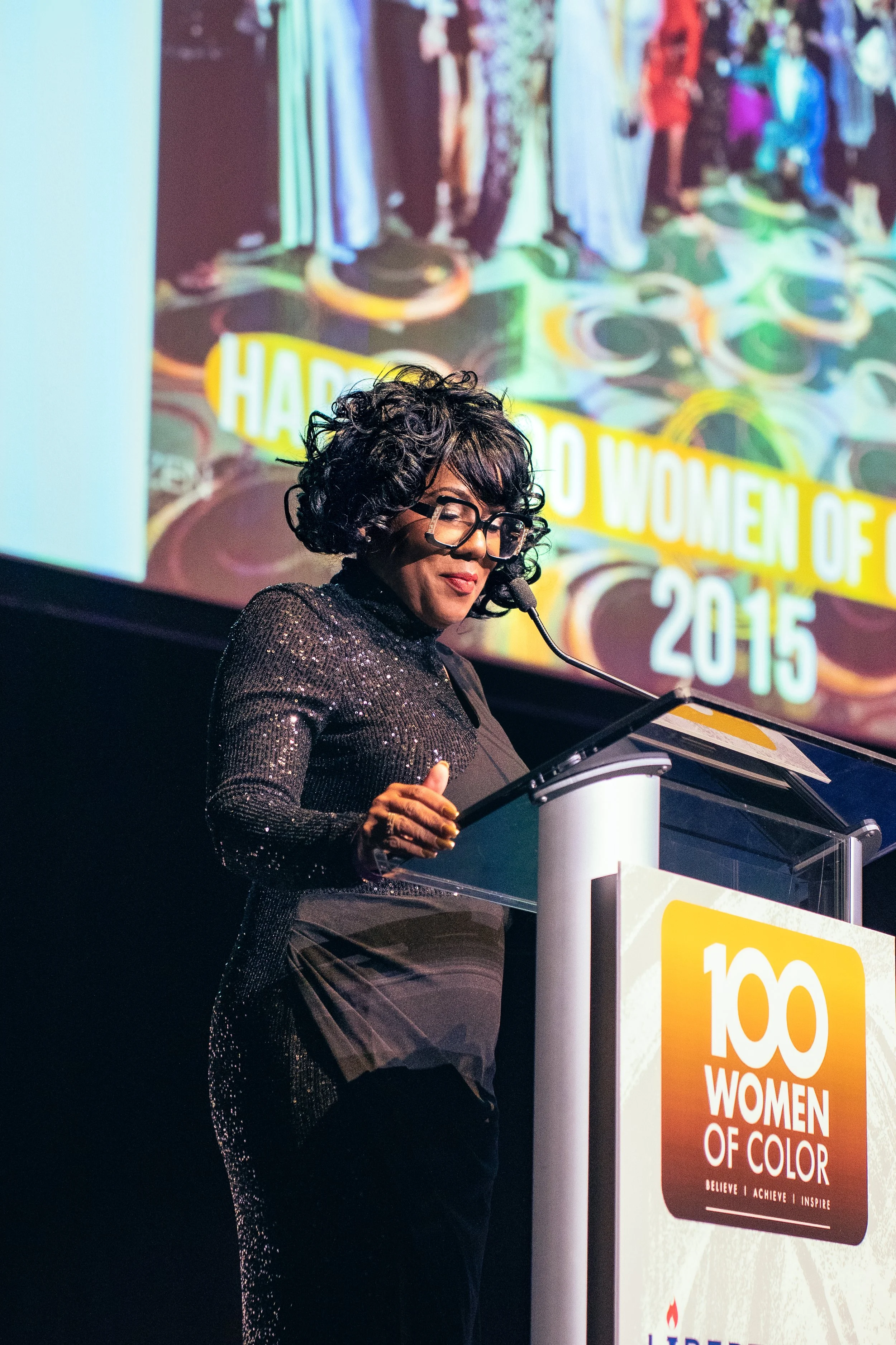 A woman with curly hair and glasses speaking at a podium during an event. The background shows a colorful banner that reads '100 Women of Color 2015' and has an array of hanging clothing items.
