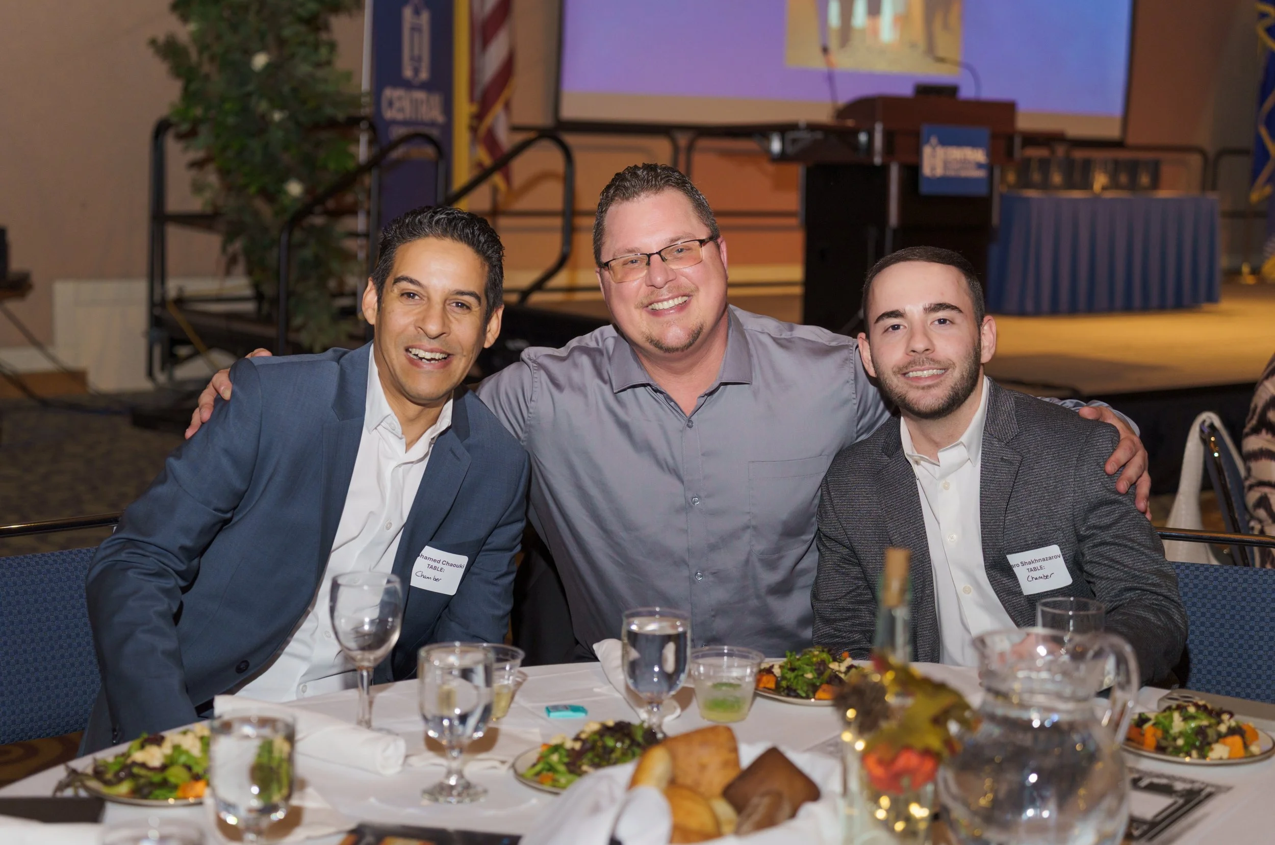 Three men sitting at a banquet table, smiling at the camera. The table is set with plates of food, wine glasses, and floral centerpieces. In the background, there is a stage with a screen and some plants.