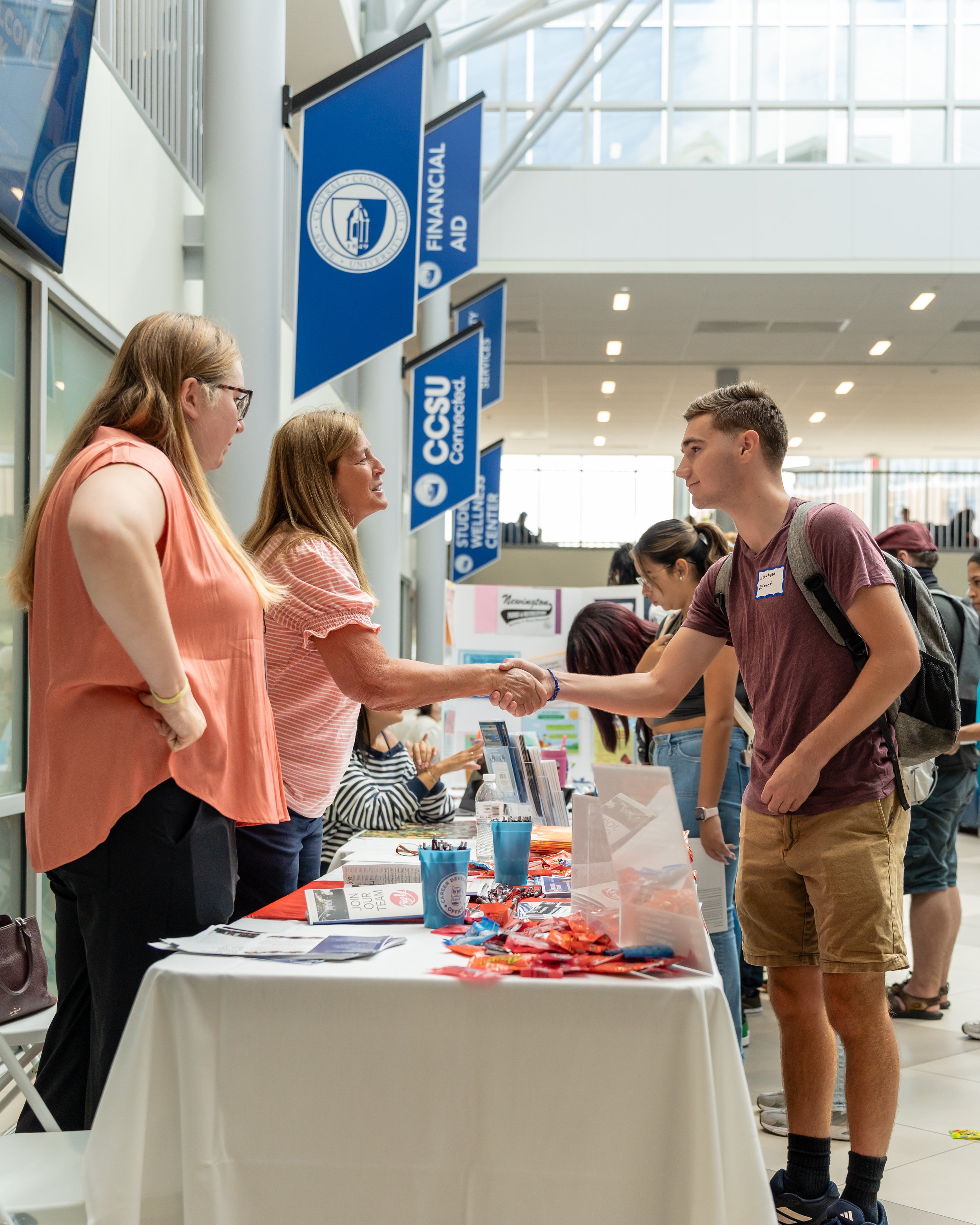 People at a university event, with a table displaying informational materials and giveaways, as students and staff engage in conversations inside a modern building with banners for the university's financial aid office hanging from the ceiling.