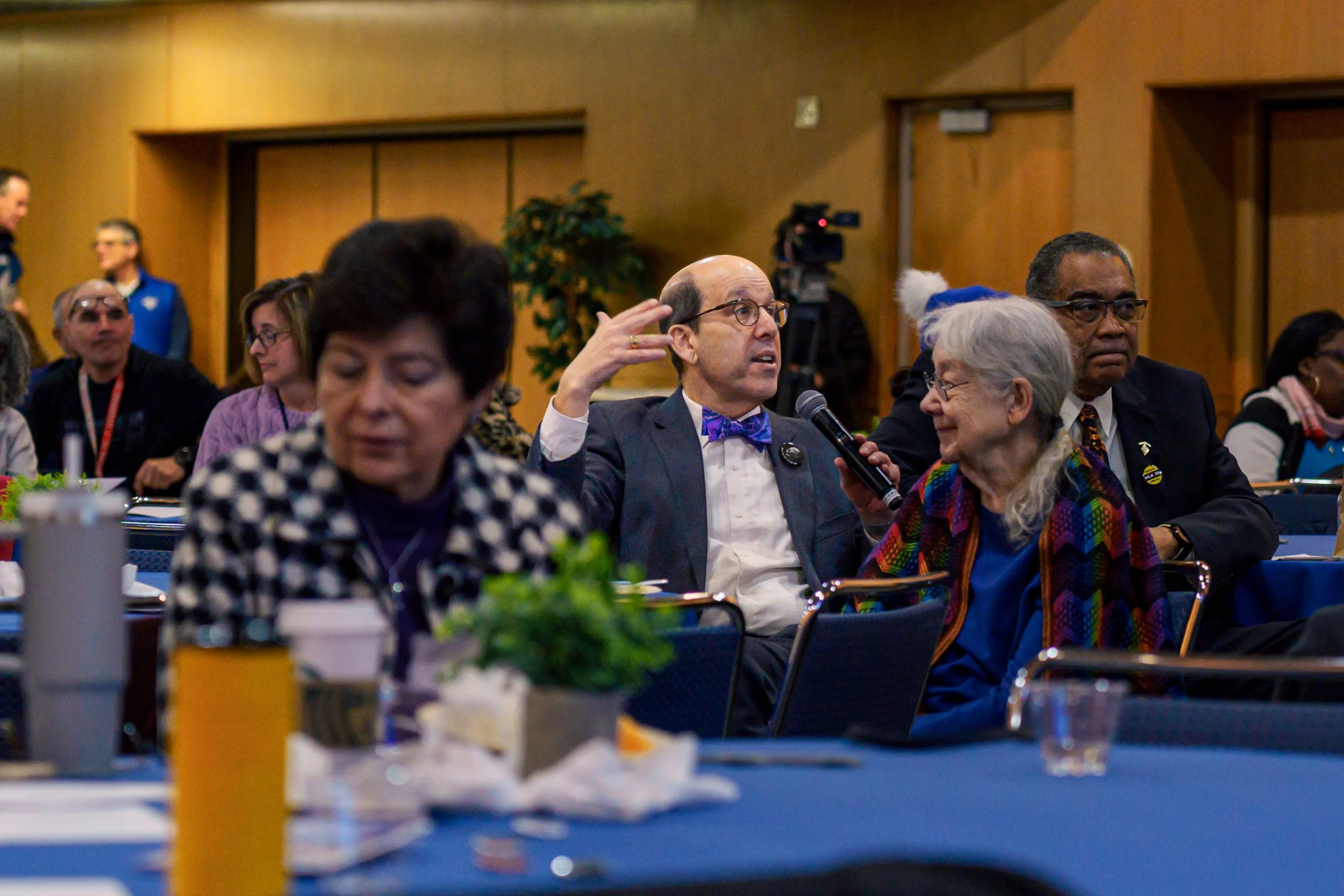 A group of people seated at a conference table, engaged in discussion; one man with glasses and a purple bow tie is speaking into a microphone, gesturing with his hand; other attendees are listening, some smiling and some looking serious. The backgro