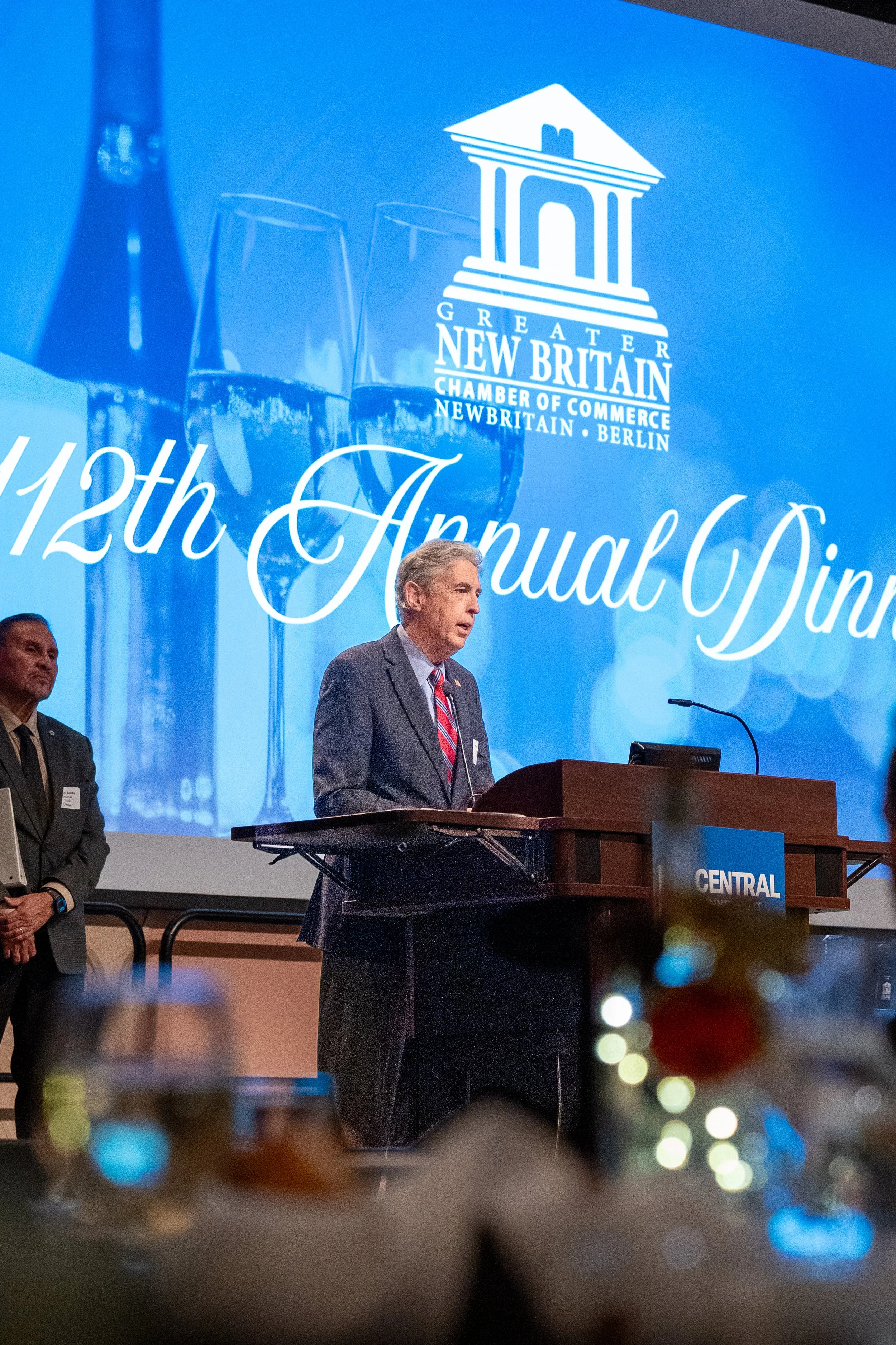 A man in a suit speaking at a podium during an event with a large blue screen behind him displaying the Great Britain Chamber of Commerce logo and the words "12th Annual Dinner."