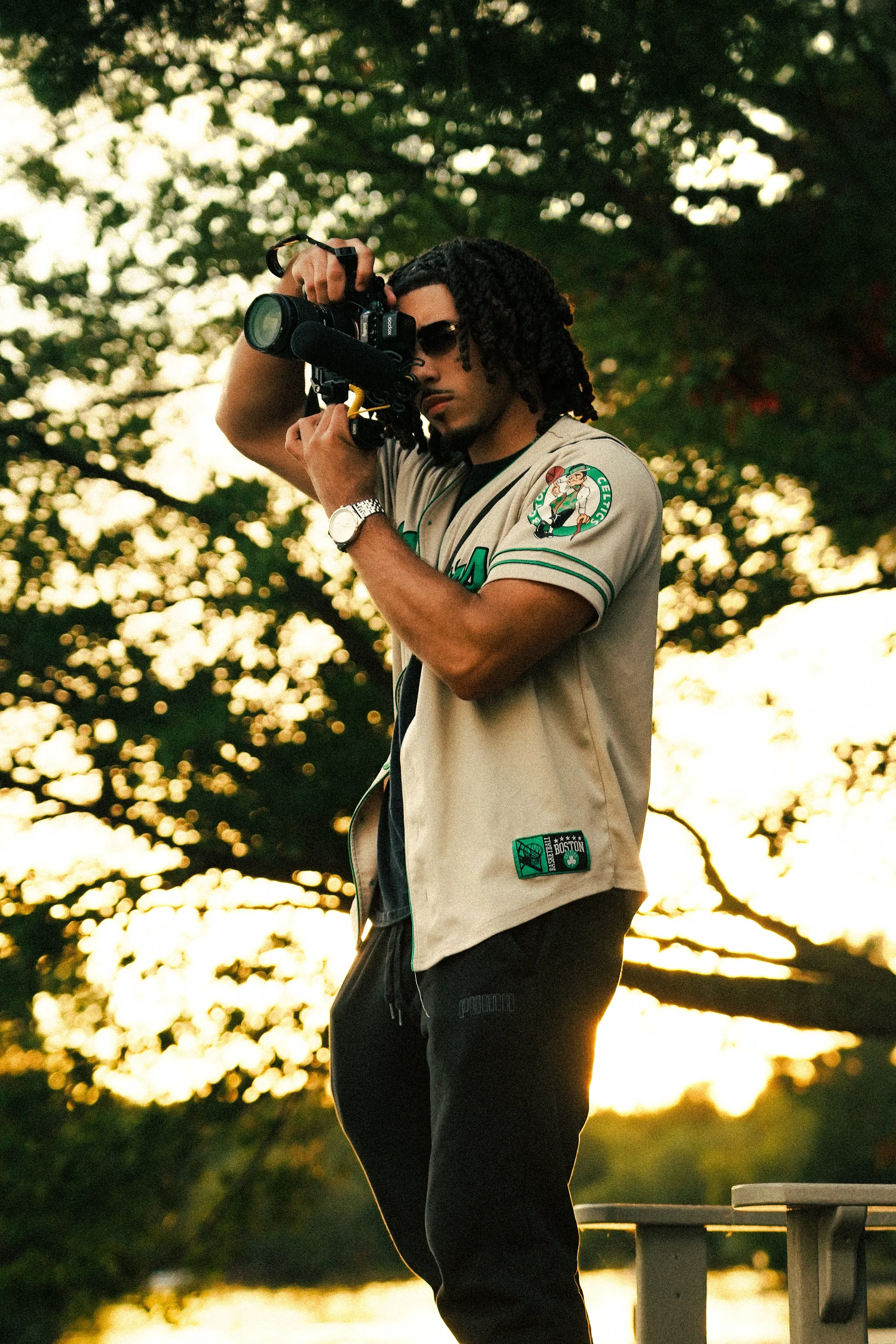 A young man with dreadlocks wearing sunglasses and a beige sports jersey with patches is filming with a camera at sunset outdoors.