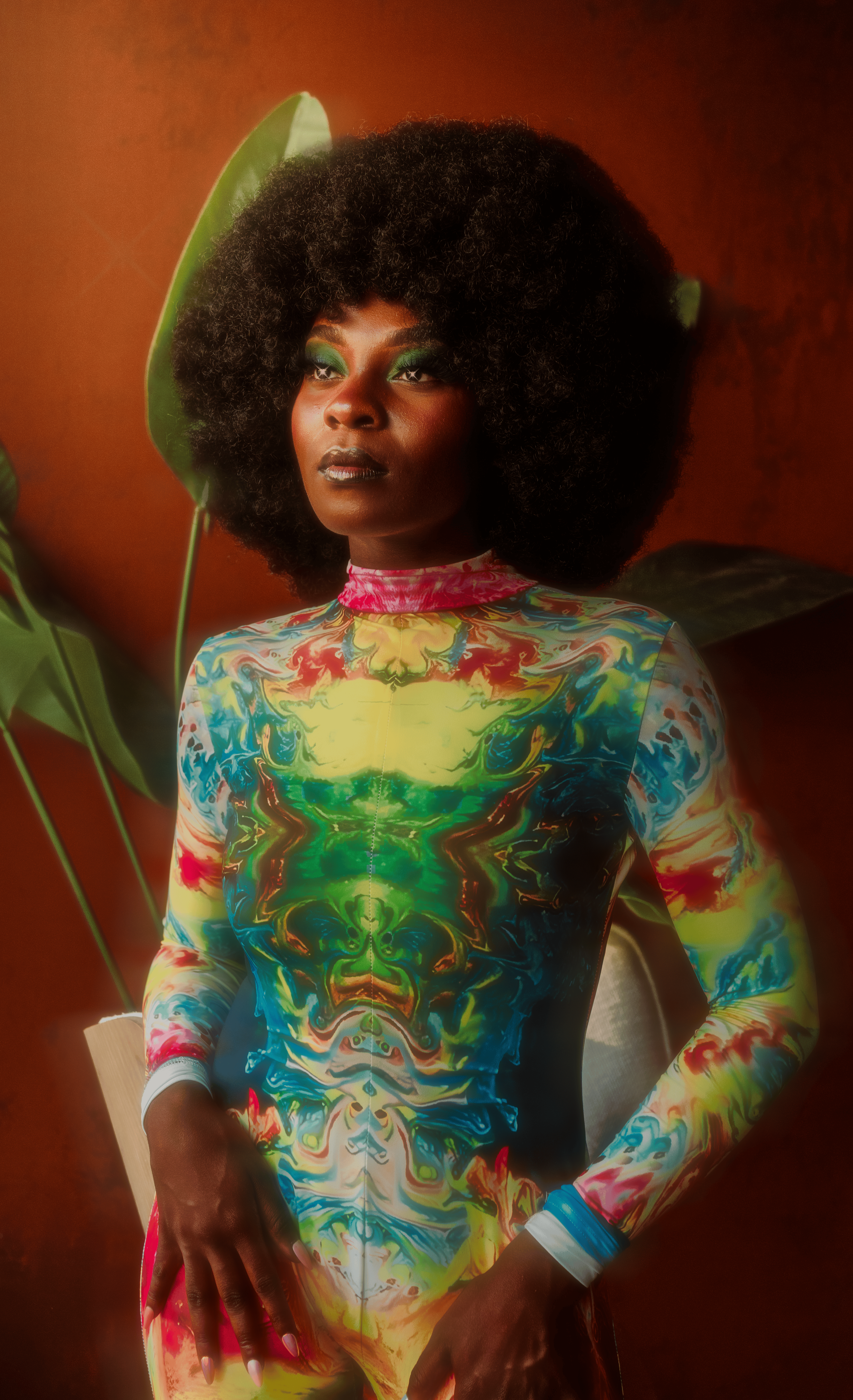 Woman with voluminous curly hair, vibrant green eye makeup, wearing a colorful abstract patterned dress, standing against a reddish background with large green leaves.